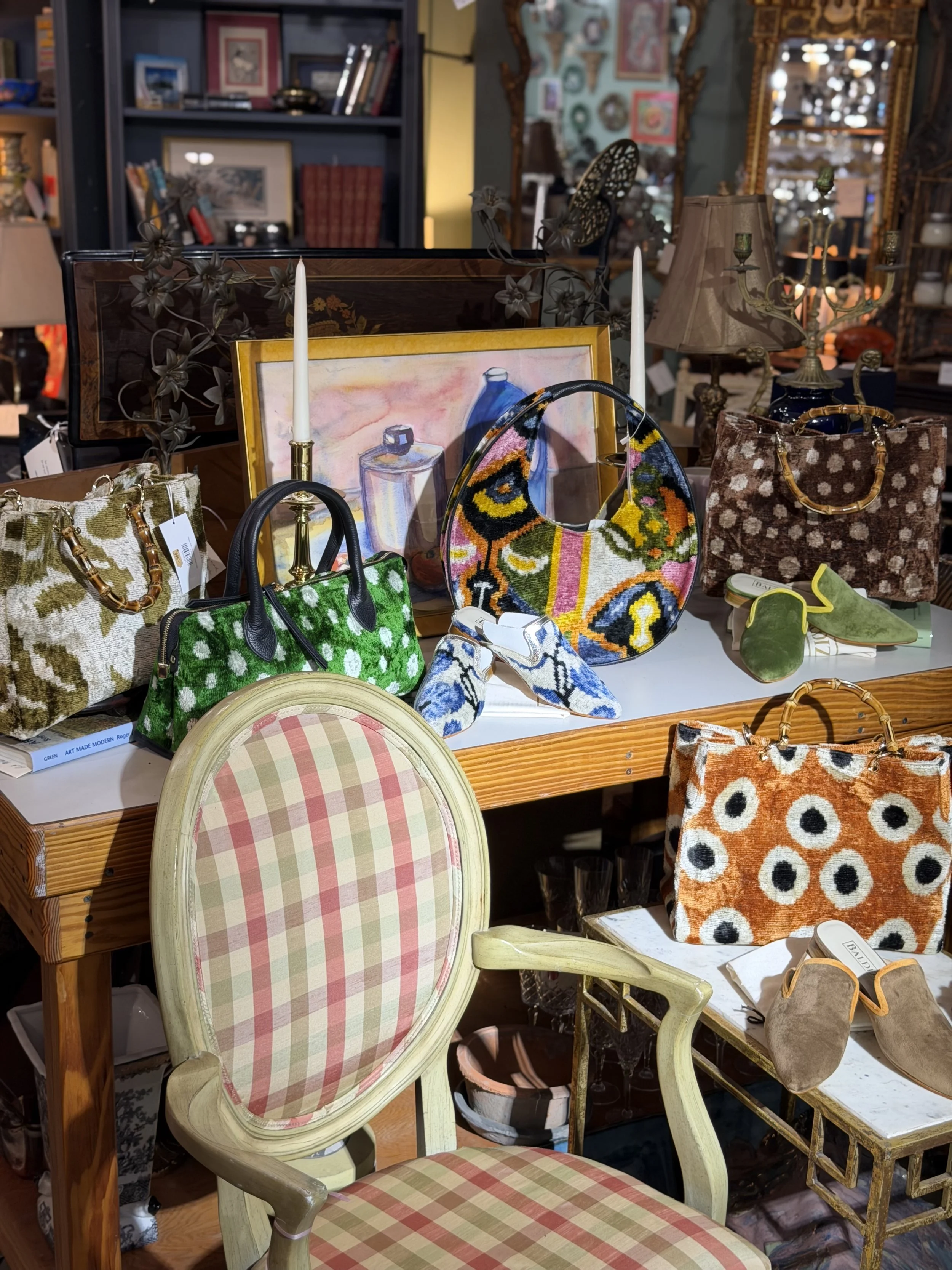 Display of colorful handbags, purses, and accessories on a table in an antique shop, with a vintage chair and framed artwork in the background.