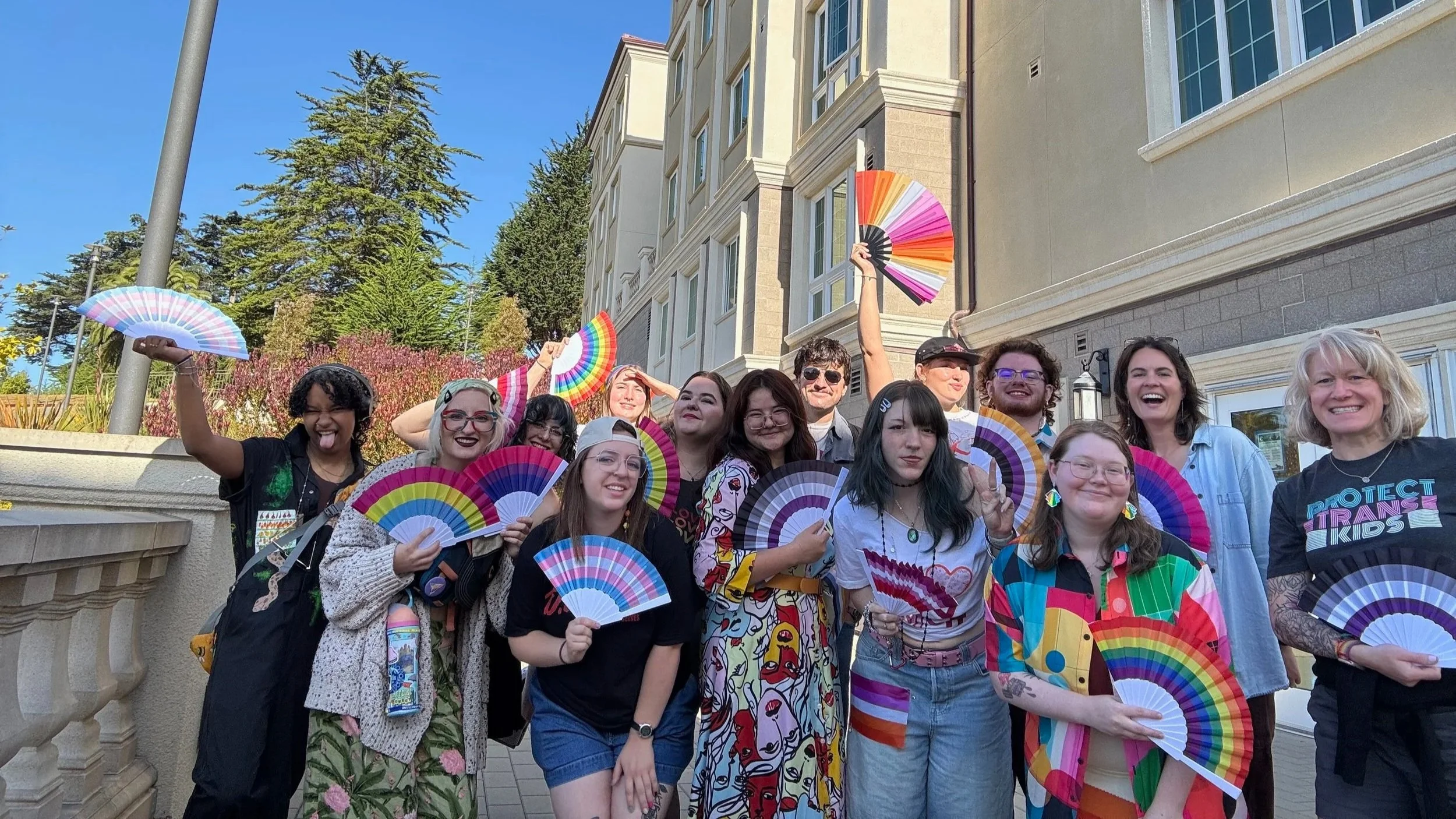 Twelve students and two program leaders stand together holding rainbow paper fans outside a brick dormitory building, smiling.