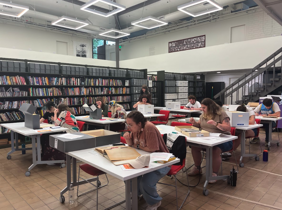 Students sit at long tables examining archival documents and gray storage boxes, with tall shelves of archive collections filling the room behind them.
