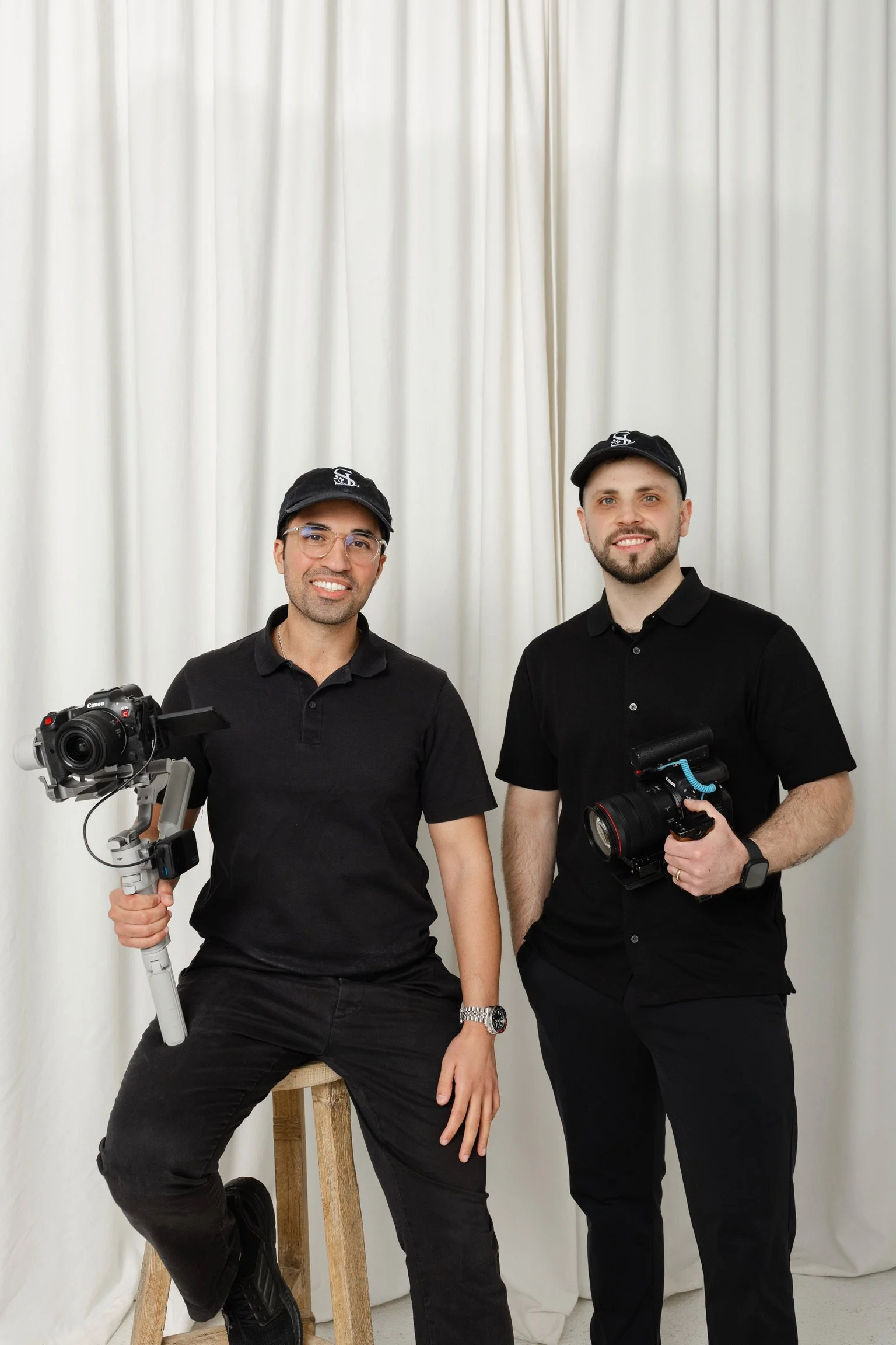 Two men in black shirts and caps holding camera equipment, standing in front of a white curtain backdrop.