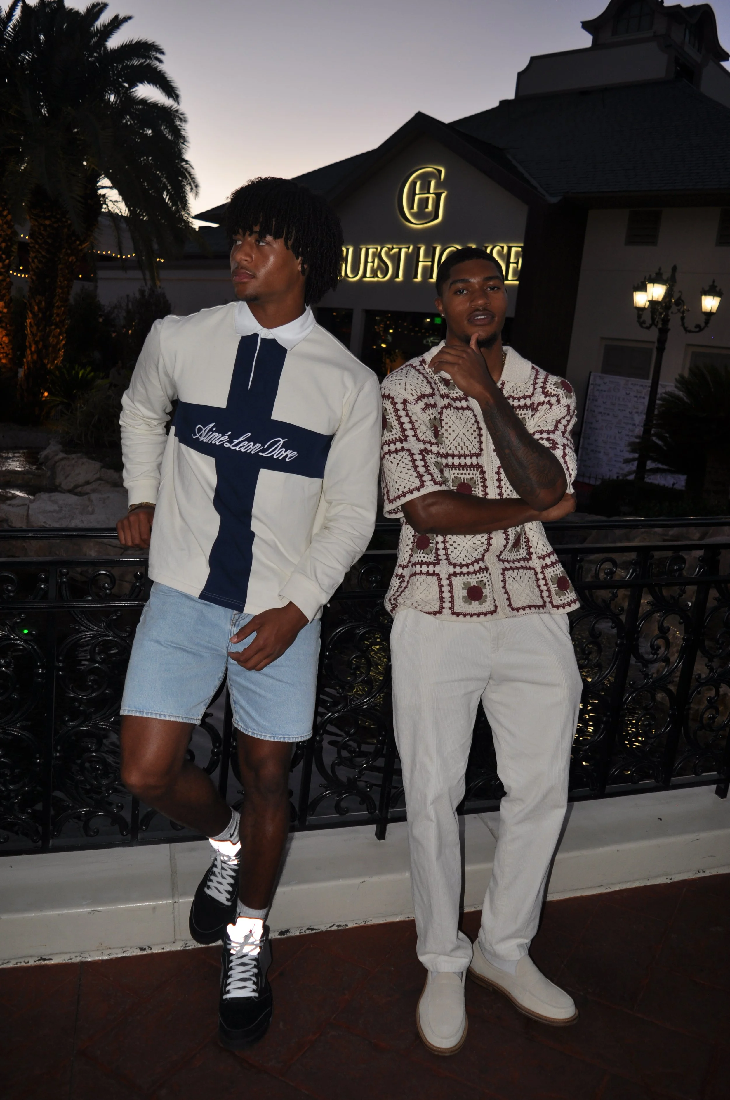 Two young men standing in front of a black wrought iron fence and a guest house sign in the evening. One is wearing a white shirt with a cross and the words 'Ainé Lean Dové', denim shorts, and black sneakers. The other is wearing a patterned short-sl