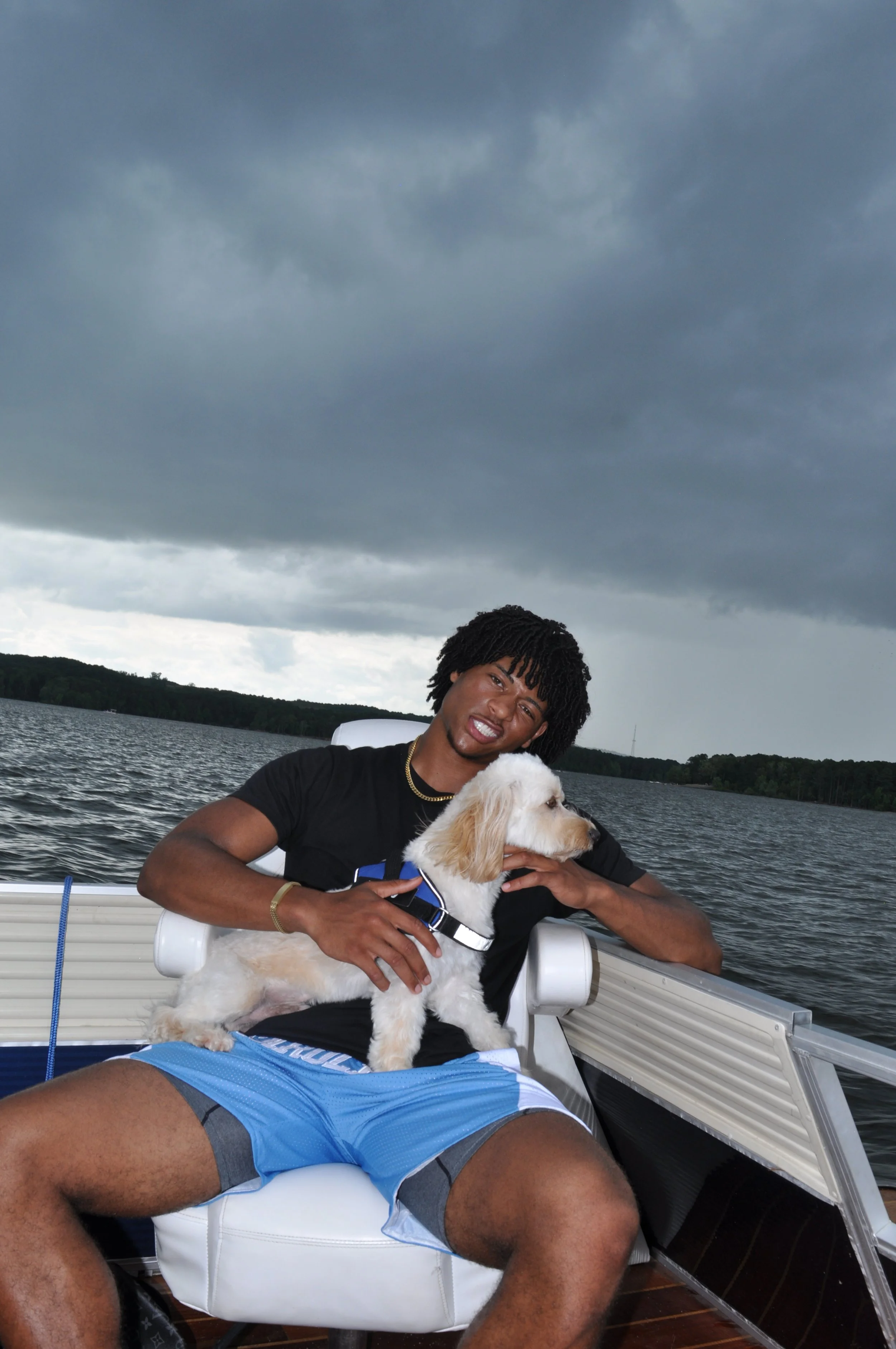 Young man sitting on a boat holding a golden retriever puppy with dark clouds overhead and water in the background.