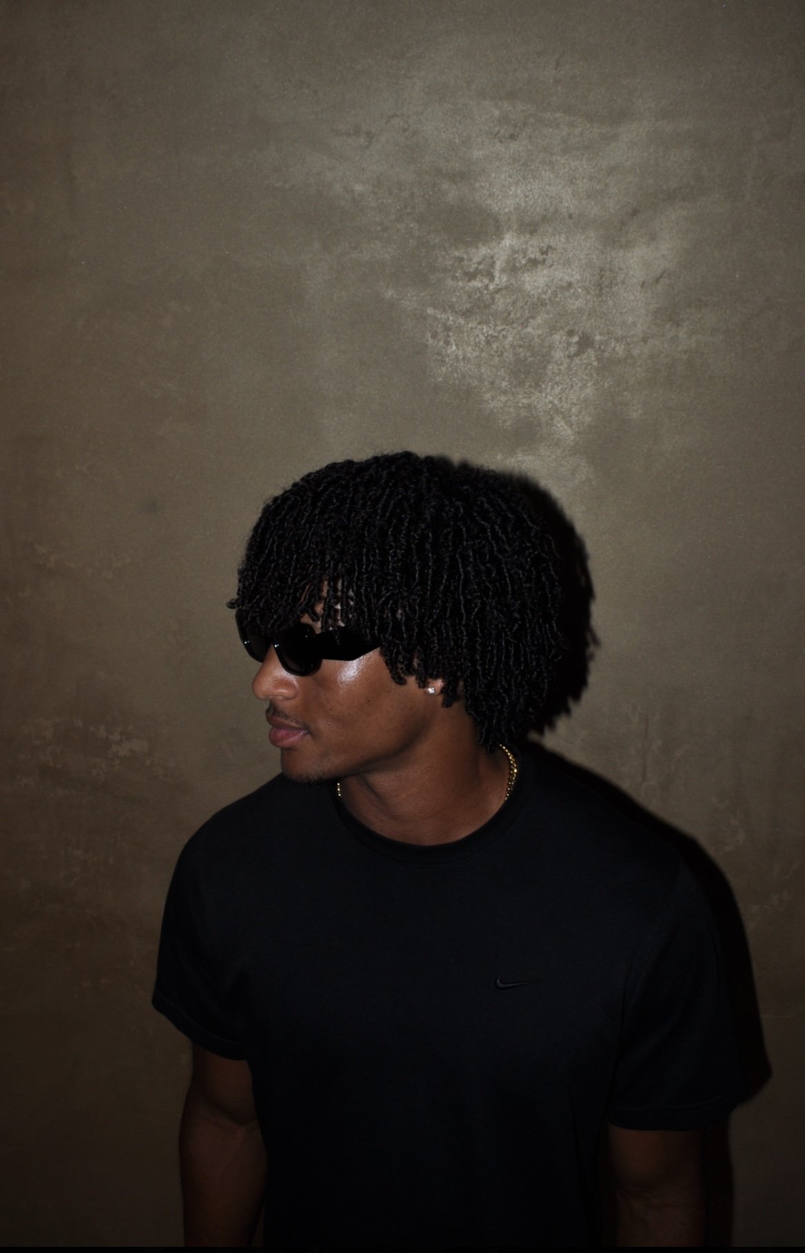 A young man wearing black sunglasses, a black Nike t-shirt, and a gold chain, standing against a textured beige wall, with his head turned to the left.