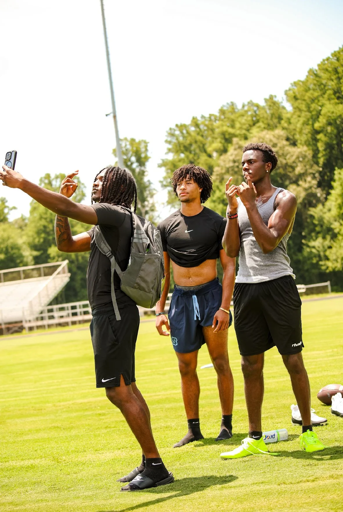 Three young men standing on a grassy field, taking a selfie or video. One is holding a smartphone, and the other two are posing thoughtfully. They are dressed in athletic clothing, with sports equipment nearby, and a background of trees and a sports 