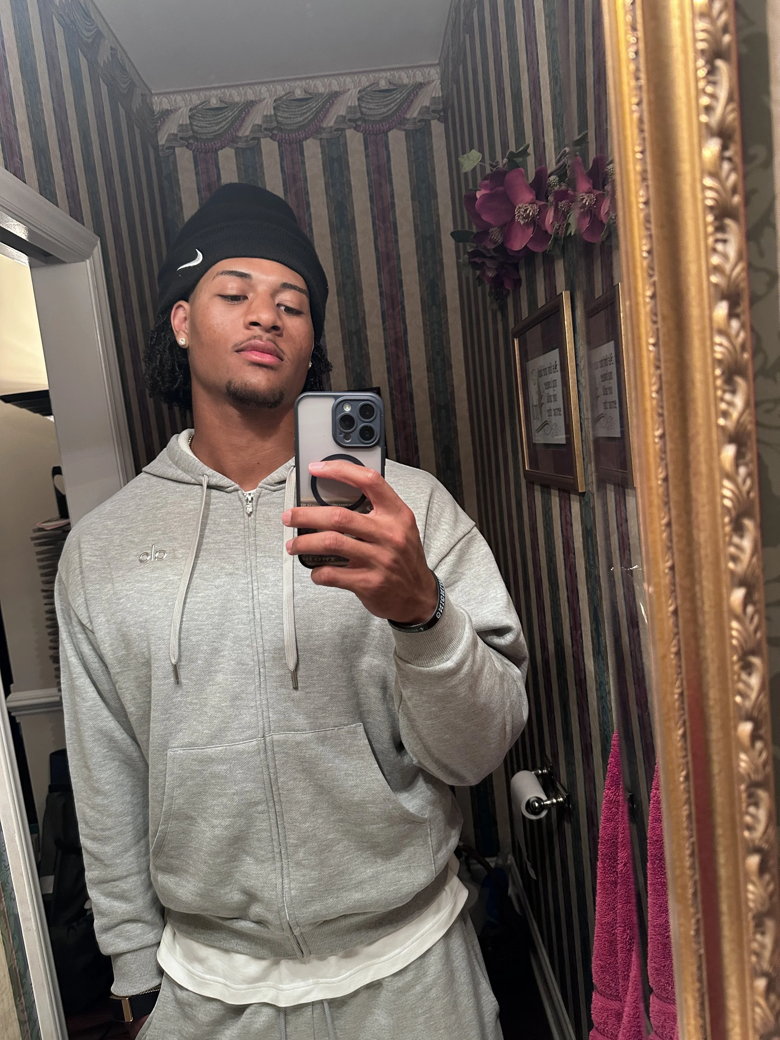 A young man taking a mirror selfie in a decorated bathroom, wearing a black beanie, gray hoodie, and gray sweatpants, with a pink towel hanging on the wall.