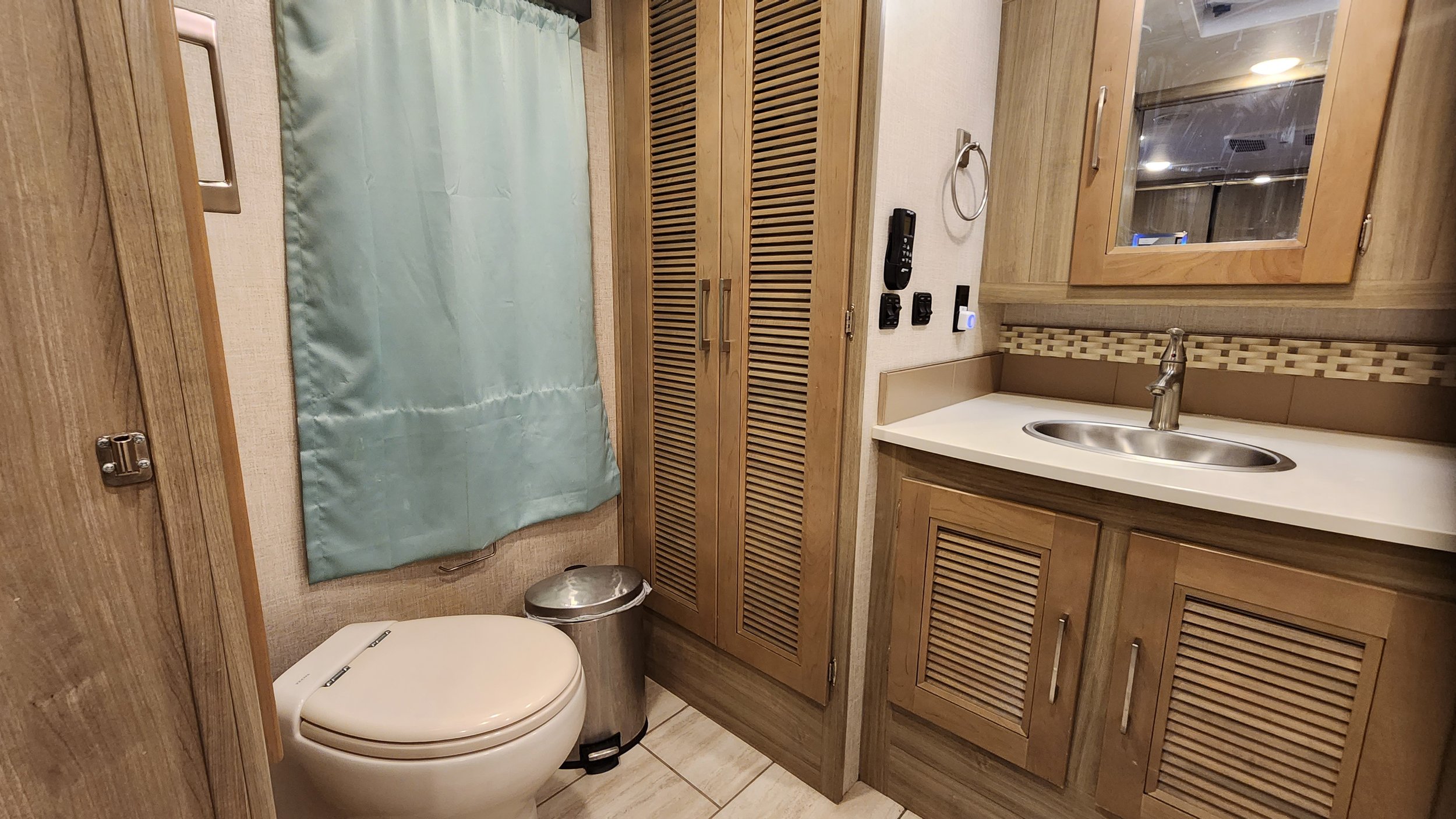 Bathroom with a toilet, a sink with a wooden cabinet, a mirror, a small window with a curtain, and a closet with louvered doors.