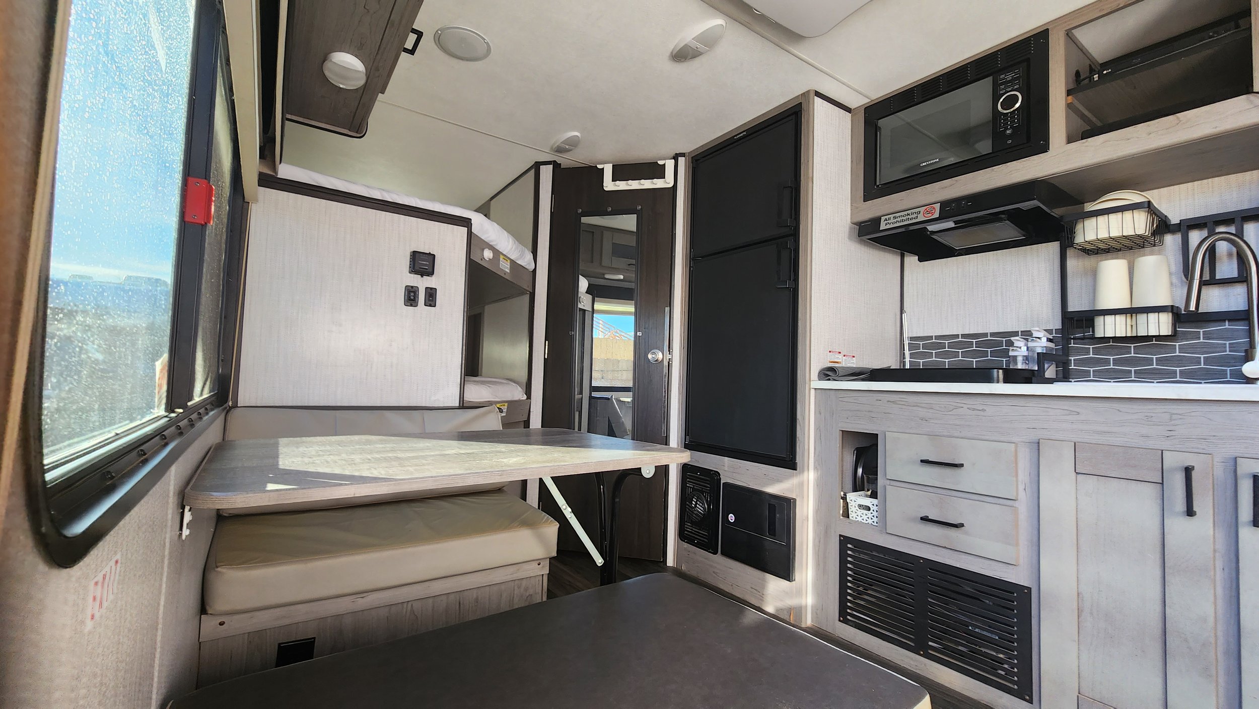 Interior of a small camper with a dining table, bunk beds, kitchenette, microwave, and small fridge.