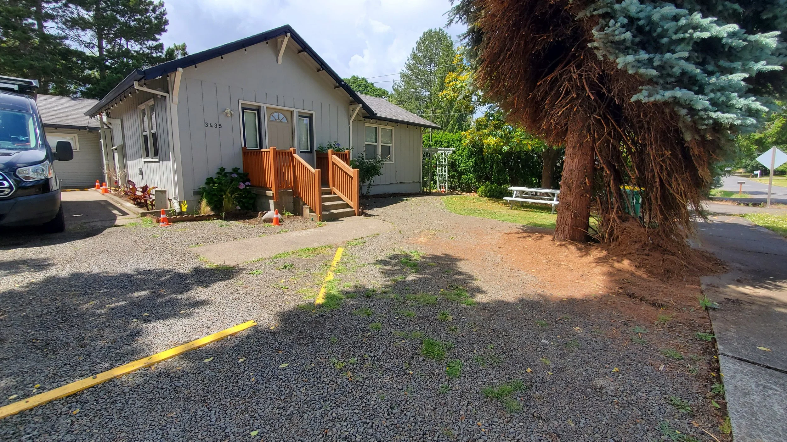 A small, single-story house with gray vertical siding, a small front porch with wooden steps and railing, and a garden bed with plants and flowers. To the left of the house is a black vehicle and a parking area with yellow parking lines. On the right