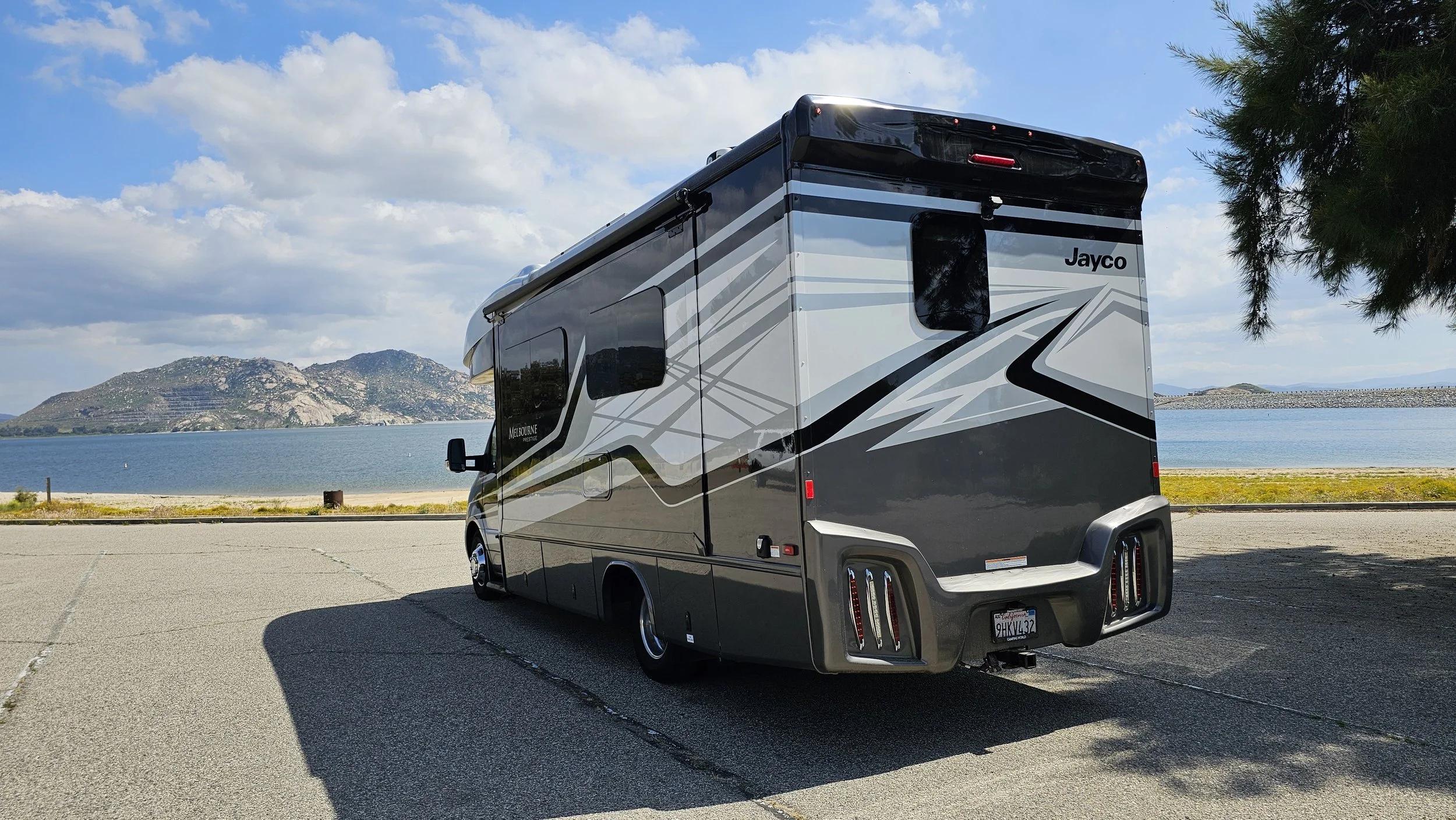 A Jayco motorhome parked near a lake with hills and partly cloudy sky in the background.