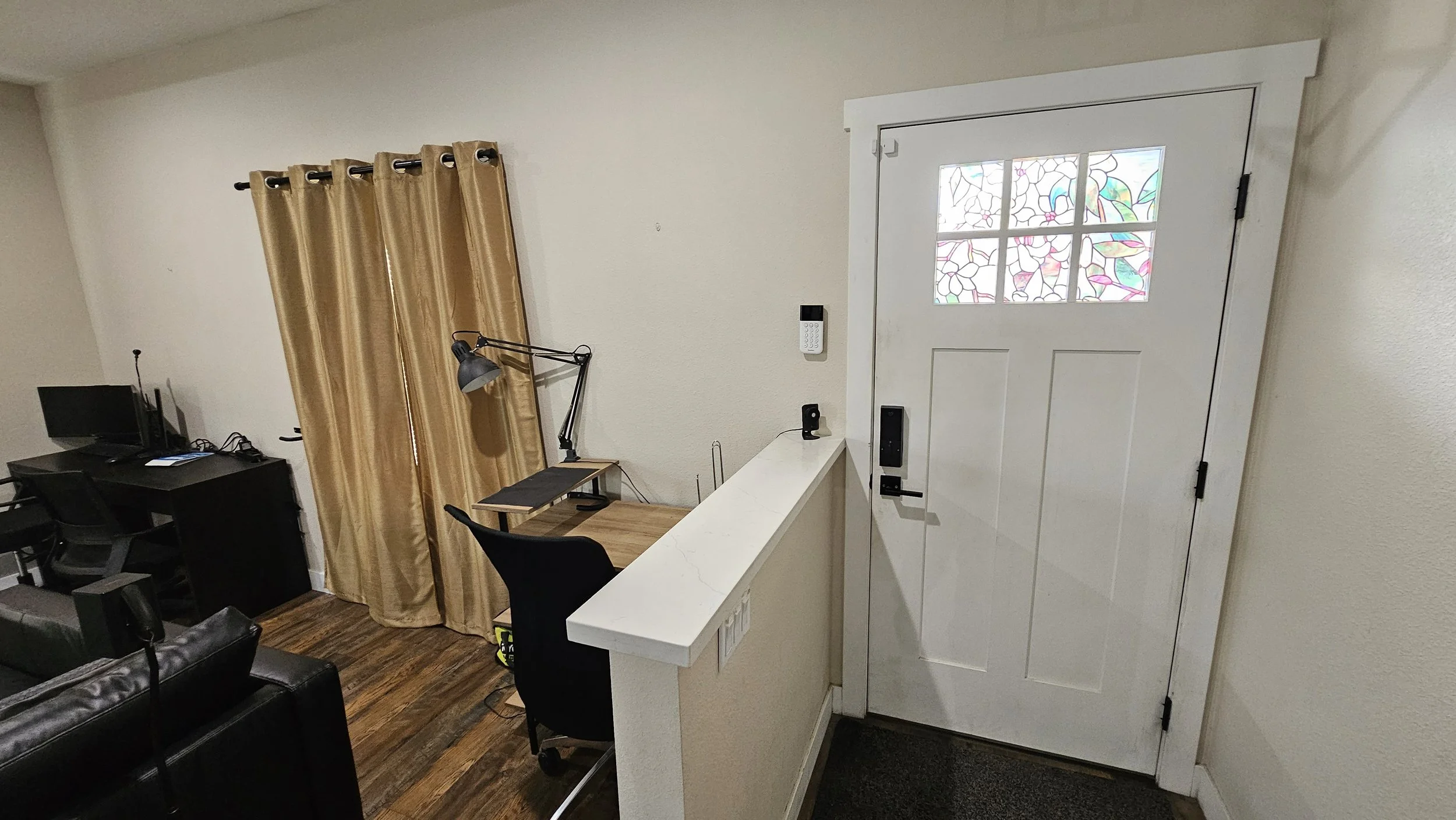 Entryway with white door featuring stained glass window, beige walls, hardwood floor, area with a black office chair, desk, floor lamp, and a gold curtain covering a window.