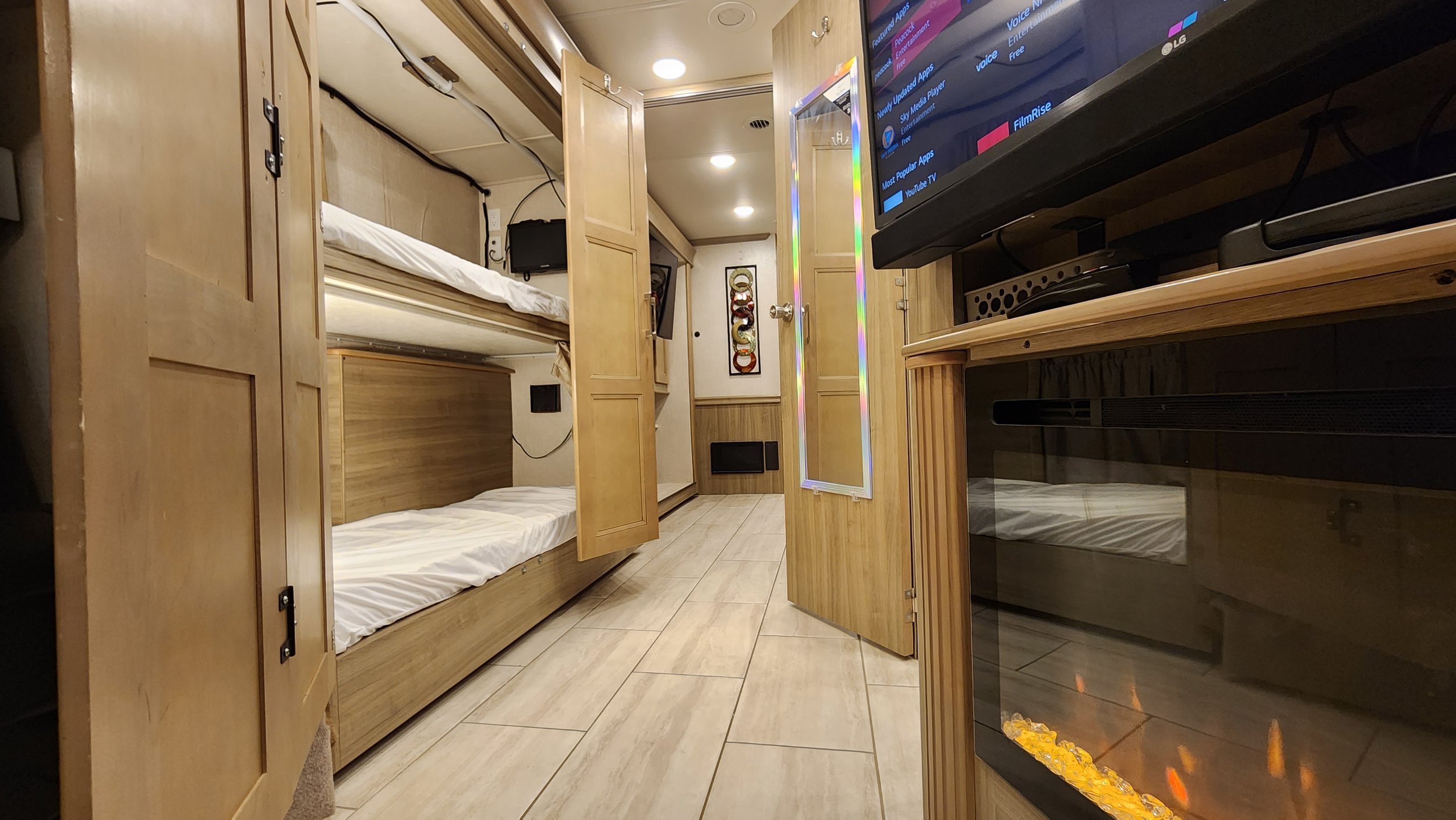 A small room with wooden bunk beds, a TV on a wooden stand, a decorative wall piece, and a mirrored door, all illuminated by recessed ceiling lights.