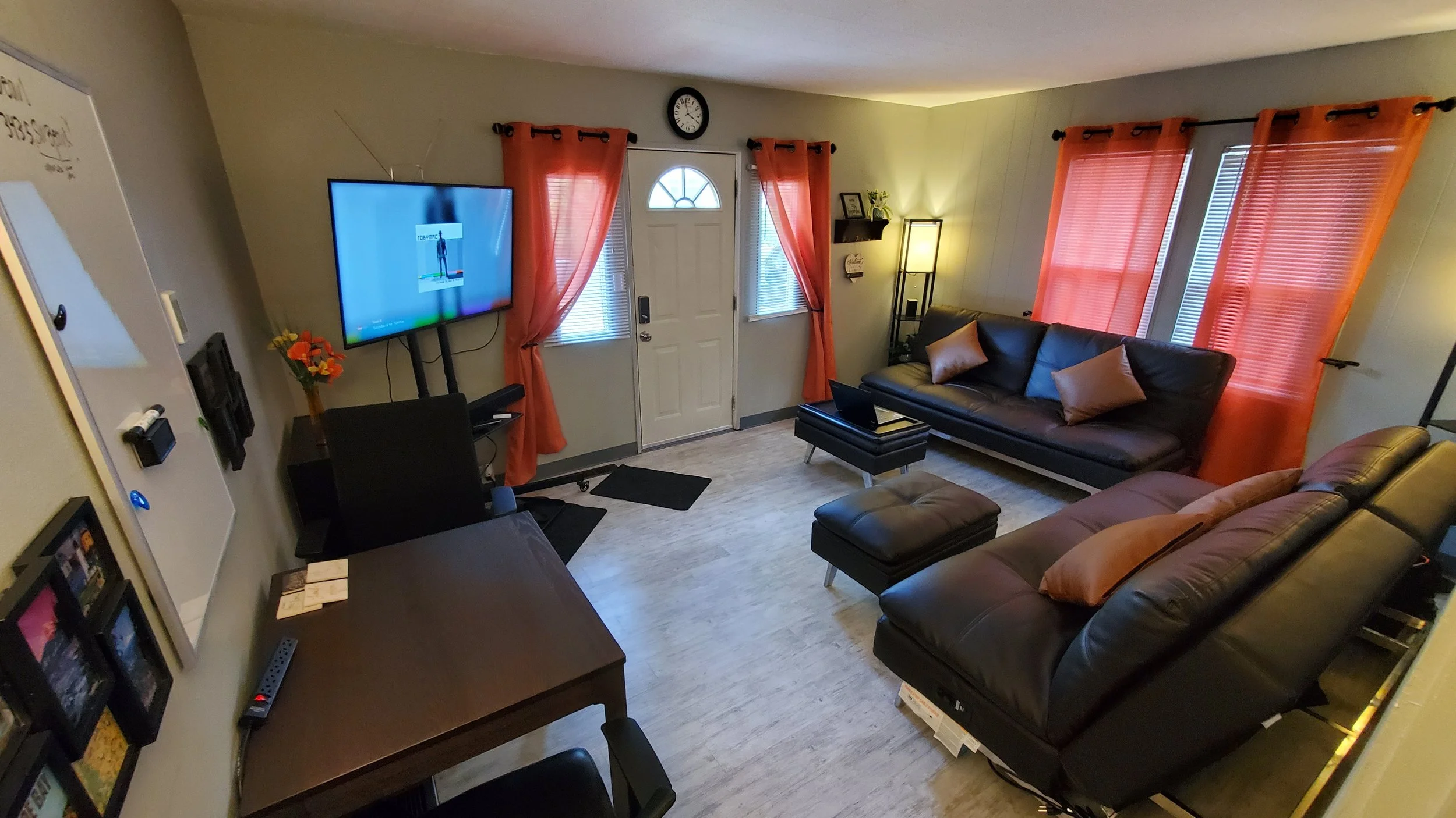 Lived-in living room with two black leather couches, a small ottoman, a flat-screen TV on a stand, an office table, and several pink curtains covering windows and door.