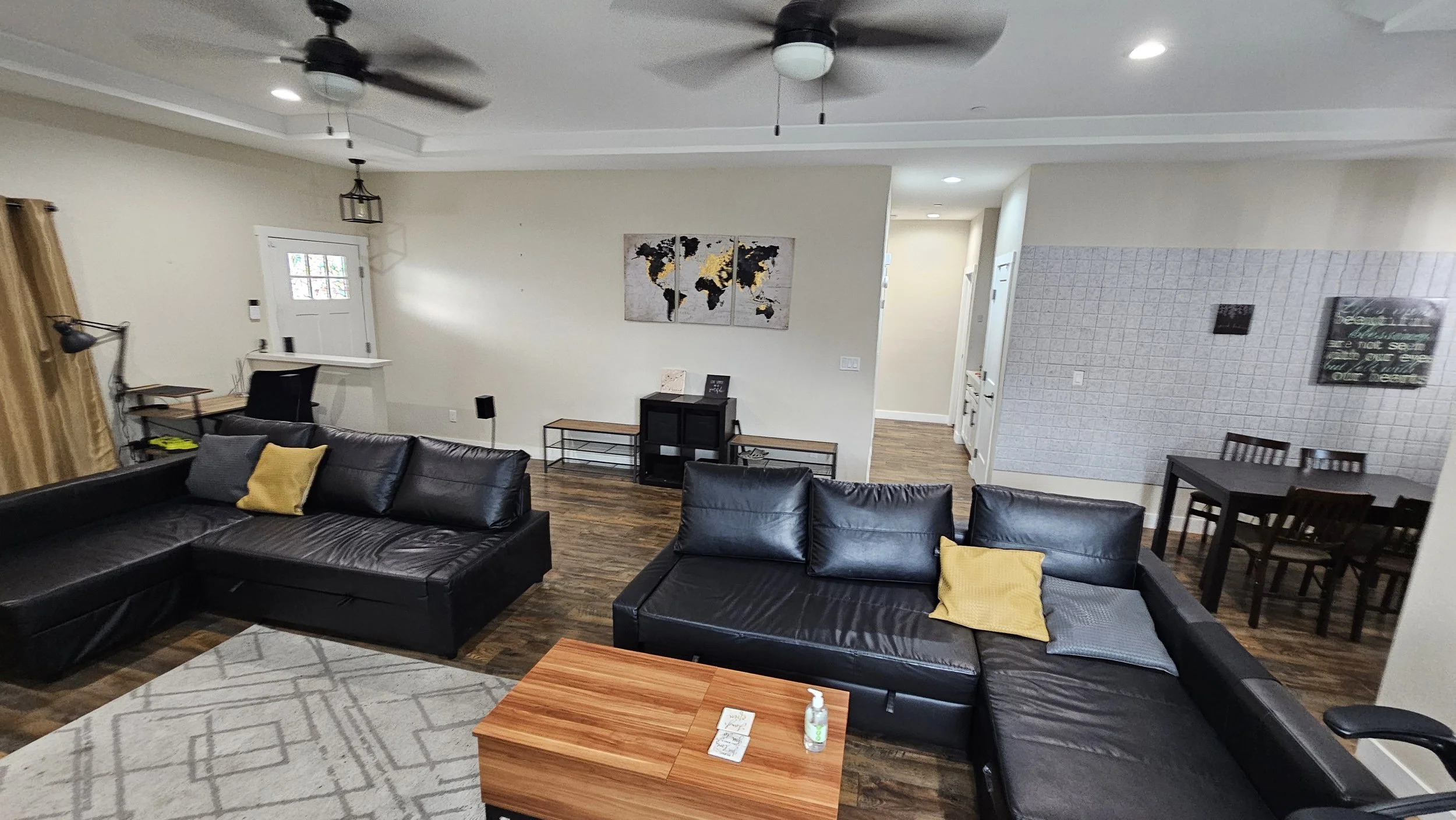 Living room with black leather sectional sofas, yellow and gray pillows, wooden coffee table, wall art of a world map, dining area with black table and chairs, and a textured wall with framed quote.
