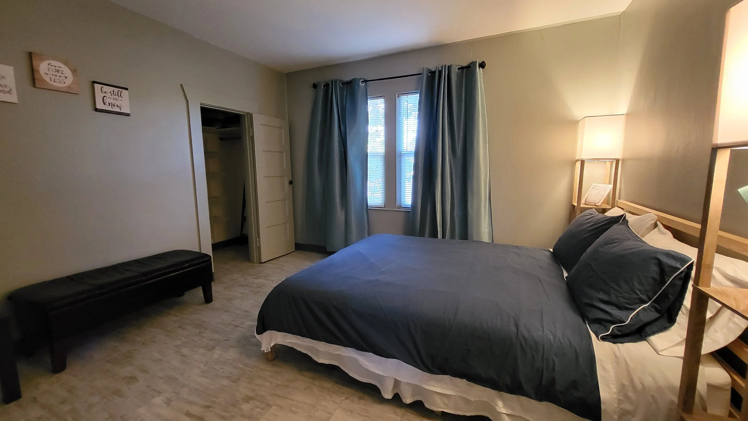 A bedroom with a bed, black bedding, pillows, a side lamp, a window with blue curtains, a bench, and wall decorations.