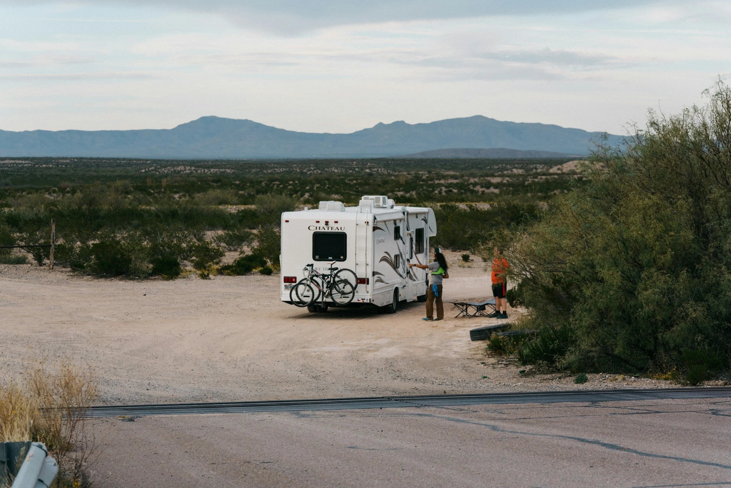 Two people standing outside a white travel trailer with bicycles attached, in a desert landscape with shrubs and mountains in the background.