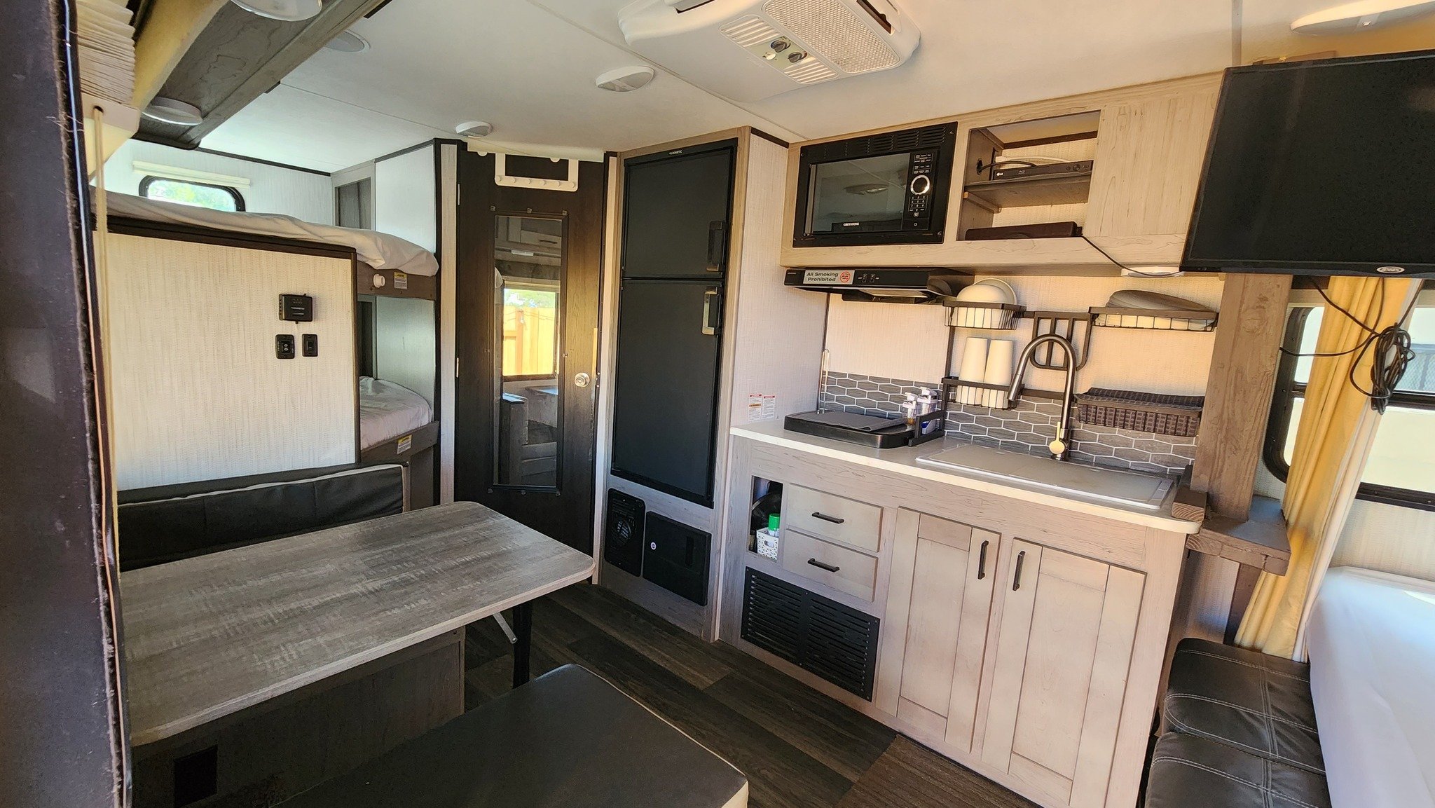 Interior of a compact living space with bunk beds, a small kitchen area with microwave, refrigerator, sink, and seating area with a table