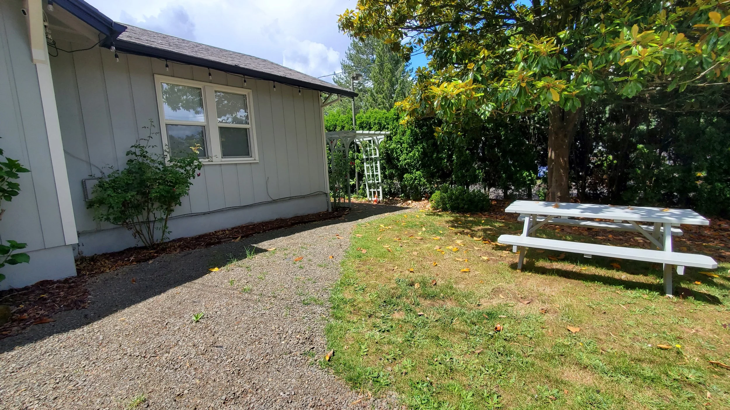 A backyard with a gravel pathway, a white house wall with windows, a white picnic table on grass, and trees and bushes in the background.