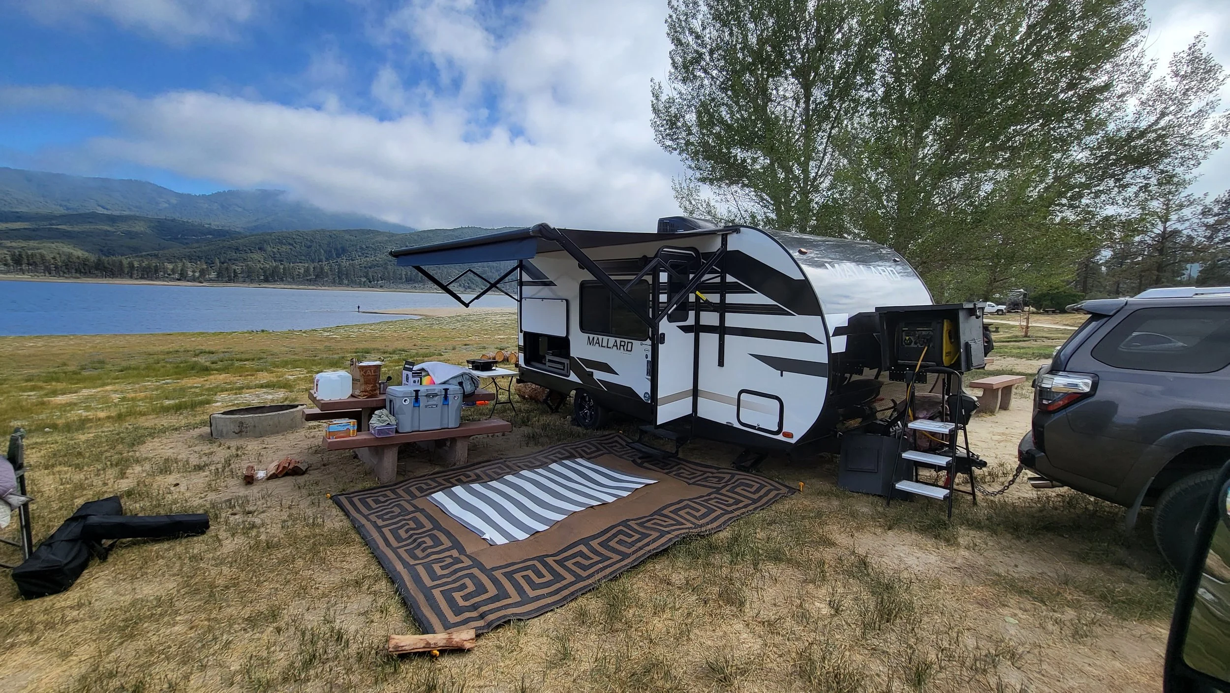 A camping trailer set up near a lake with gear and supplies on a picnic table, a gray SUV parked nearby, and a scenic backdrop of mountains, trees, and a partly cloudy sky.