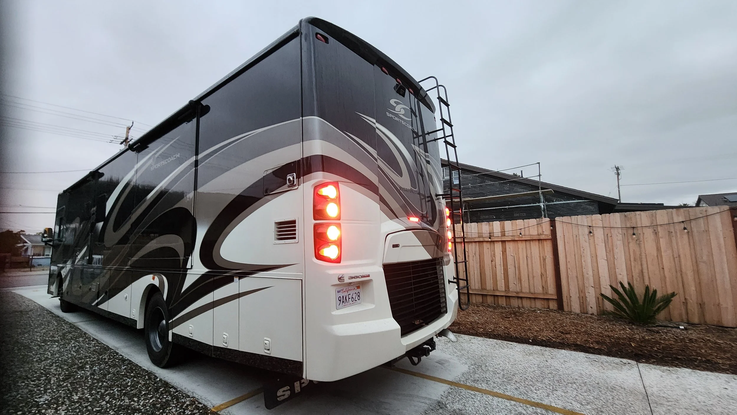 Black and white sports coach RV parked on a driveway with a wooden fence in the background and cloudy sky overhead.