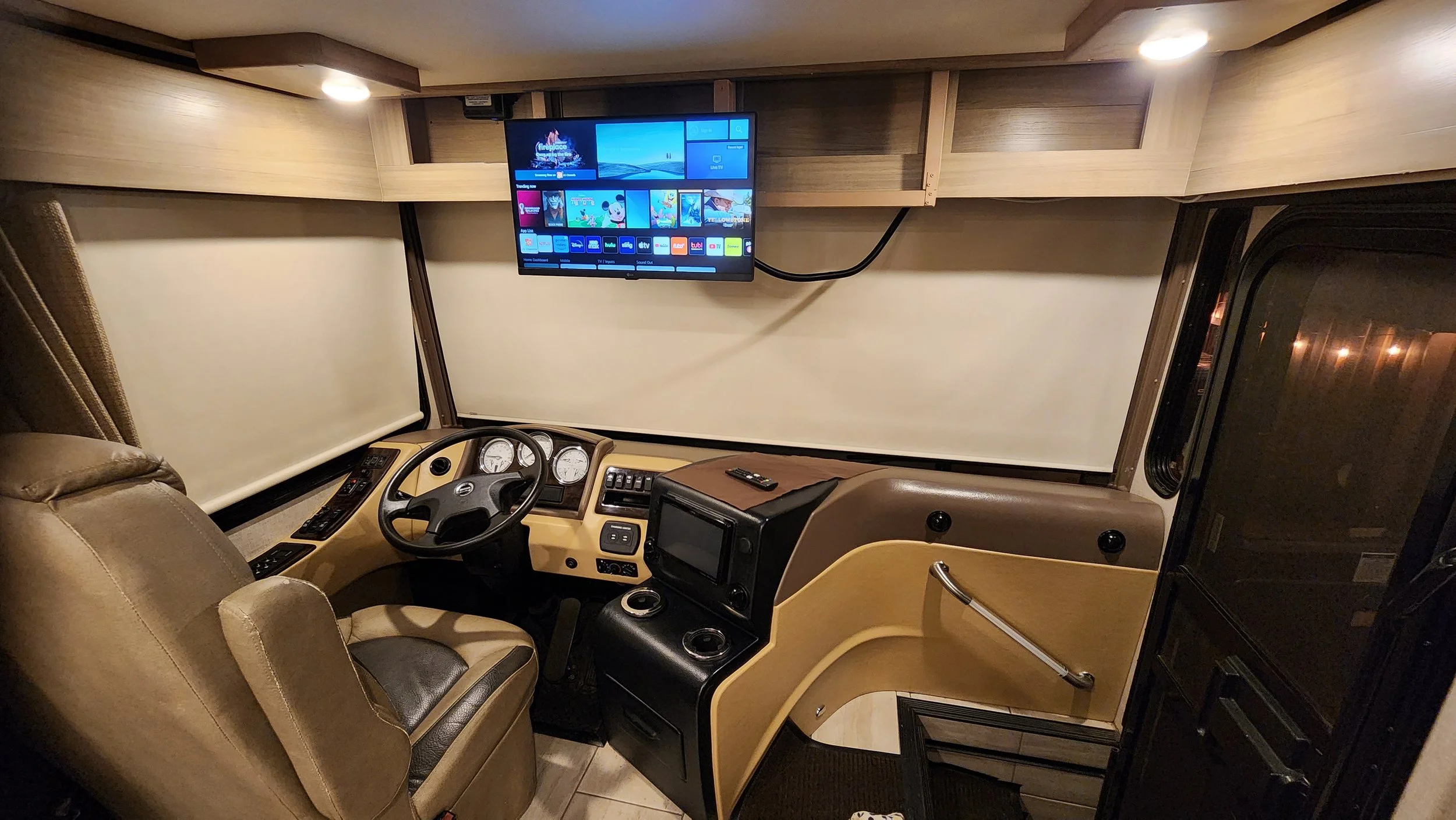 Interior of a recreational vehicle dashboard with a steering wheel, control panel, remote, and a mounted flat-screen TV.
