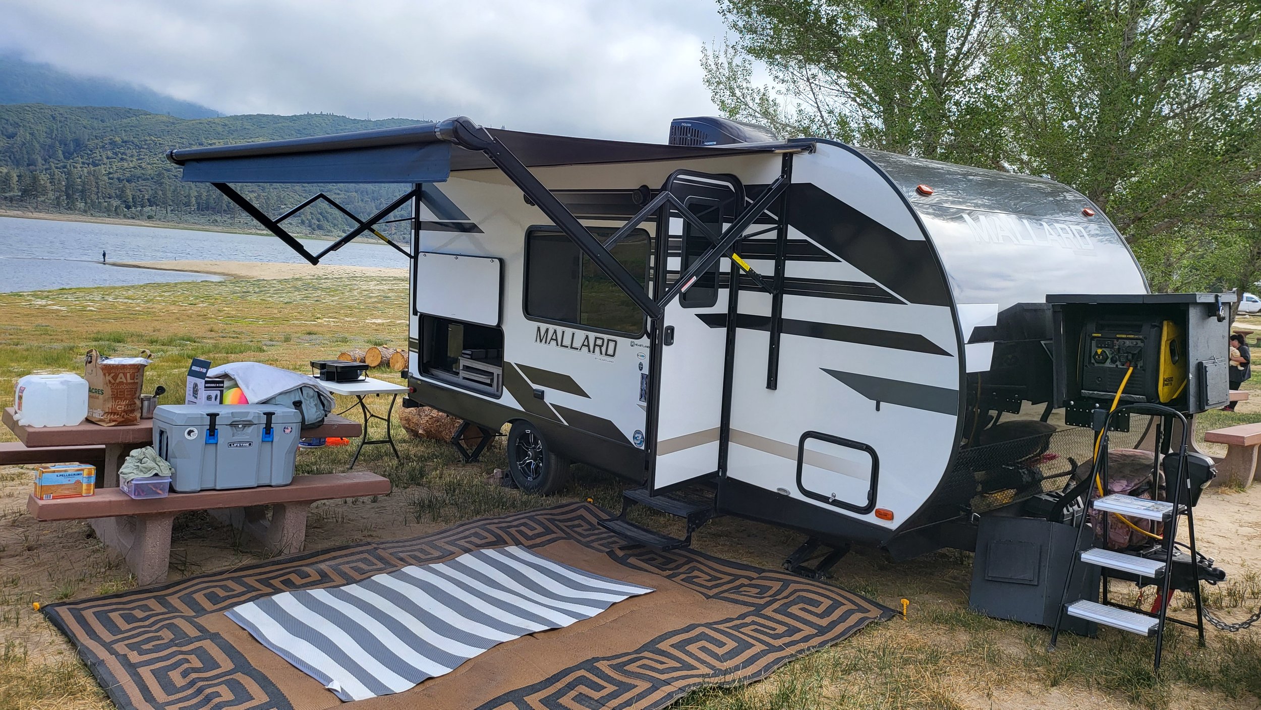 Travel trailer with outdoor setup by a lake, picnic tables, outdoor rugs, and camping gear on a grassy area with trees and mountains in the background.