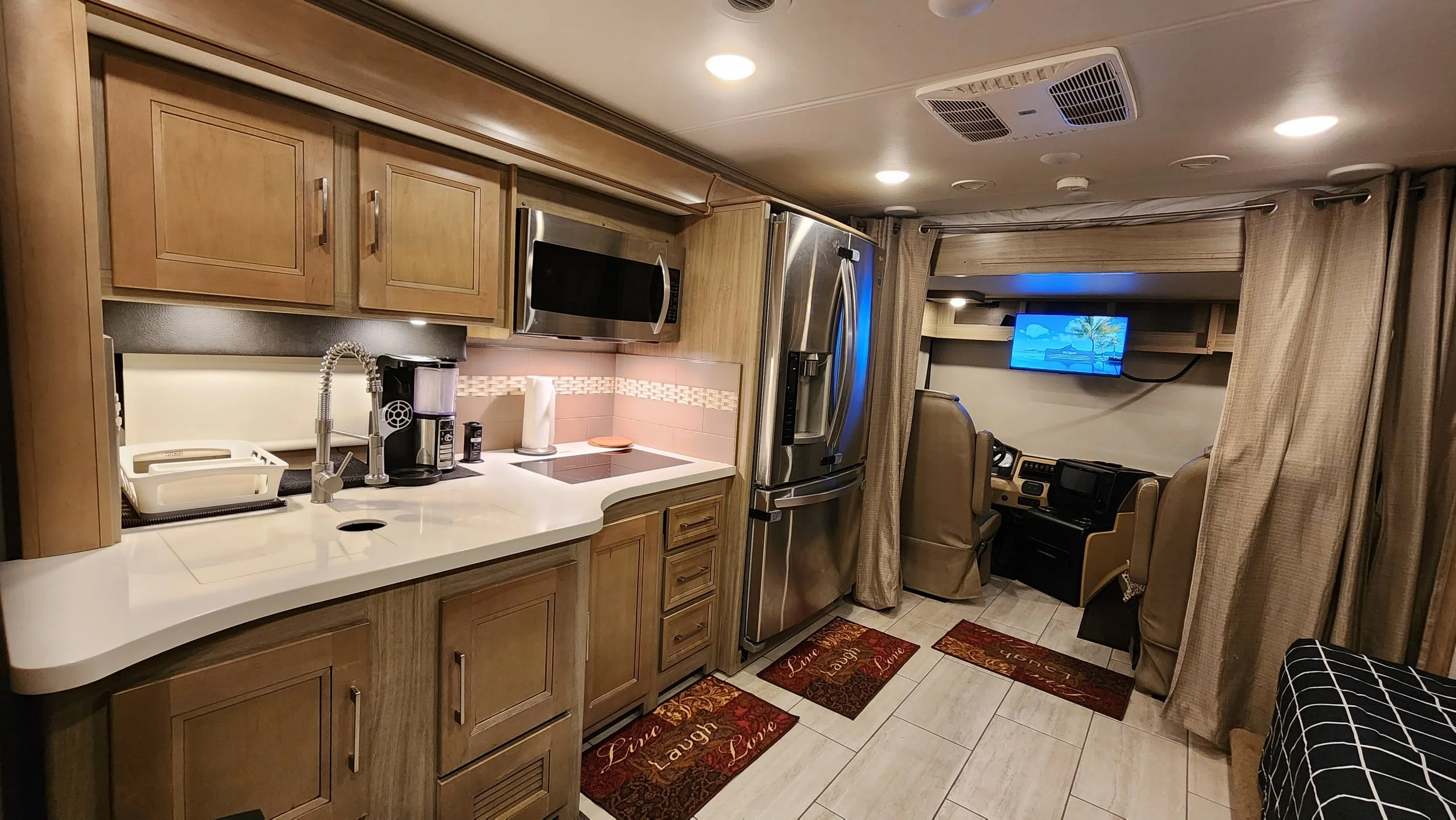 Kitchen area with wooden cabinets, stainless steel refrigerator, microwave, coffee maker, and sink, with a view into a living space with a TV and two chairs.