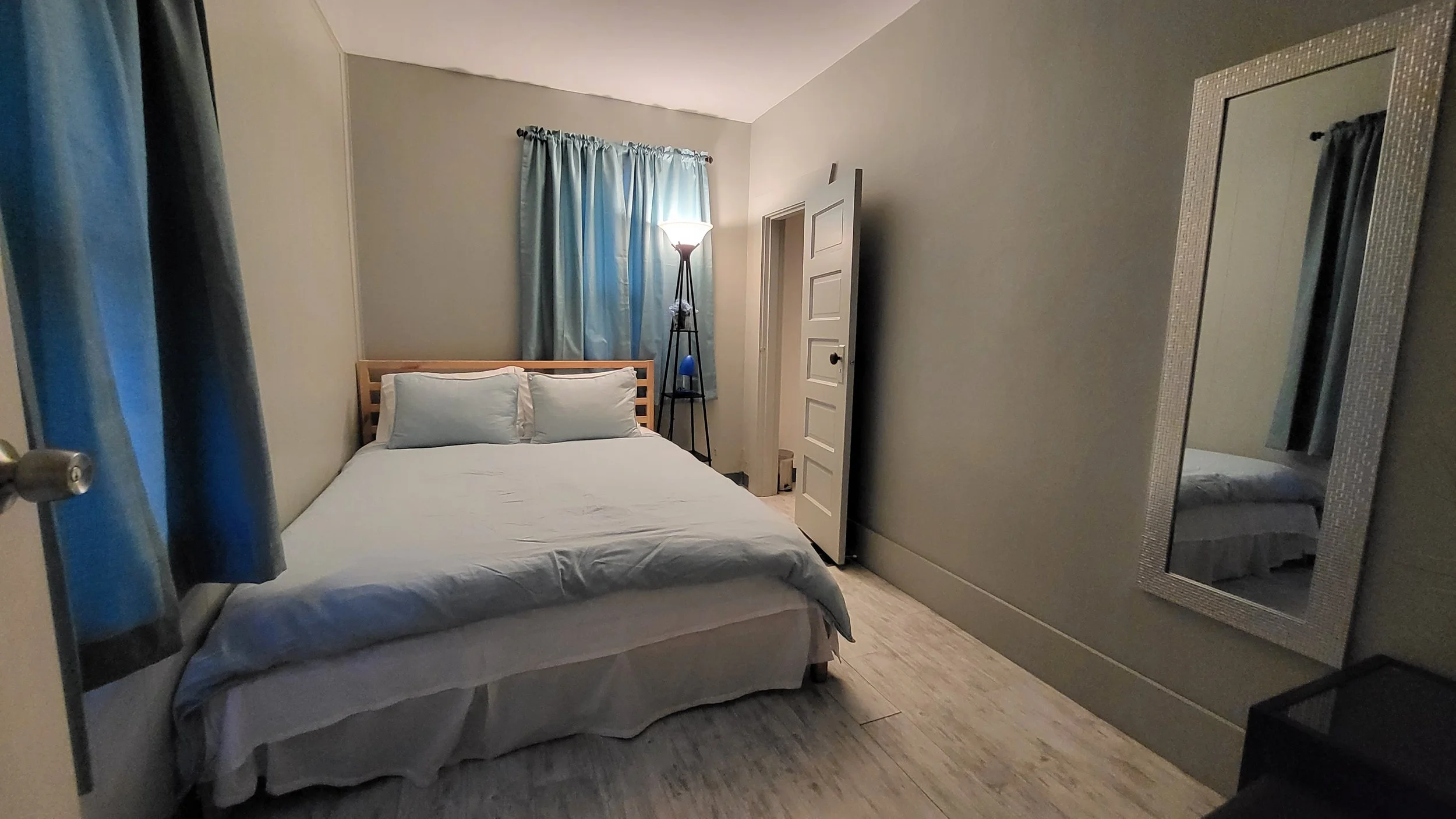 A small bedroom featuring a bed with white bedding and two pillows, a tall floor lamp beside the bed, a mirror on the wall, and a window with blue curtains.