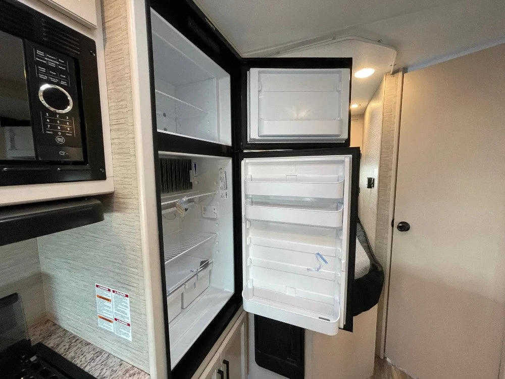 Open empty refrigerator in a kitchen next to a microwave, with a beige wall and door in the background.