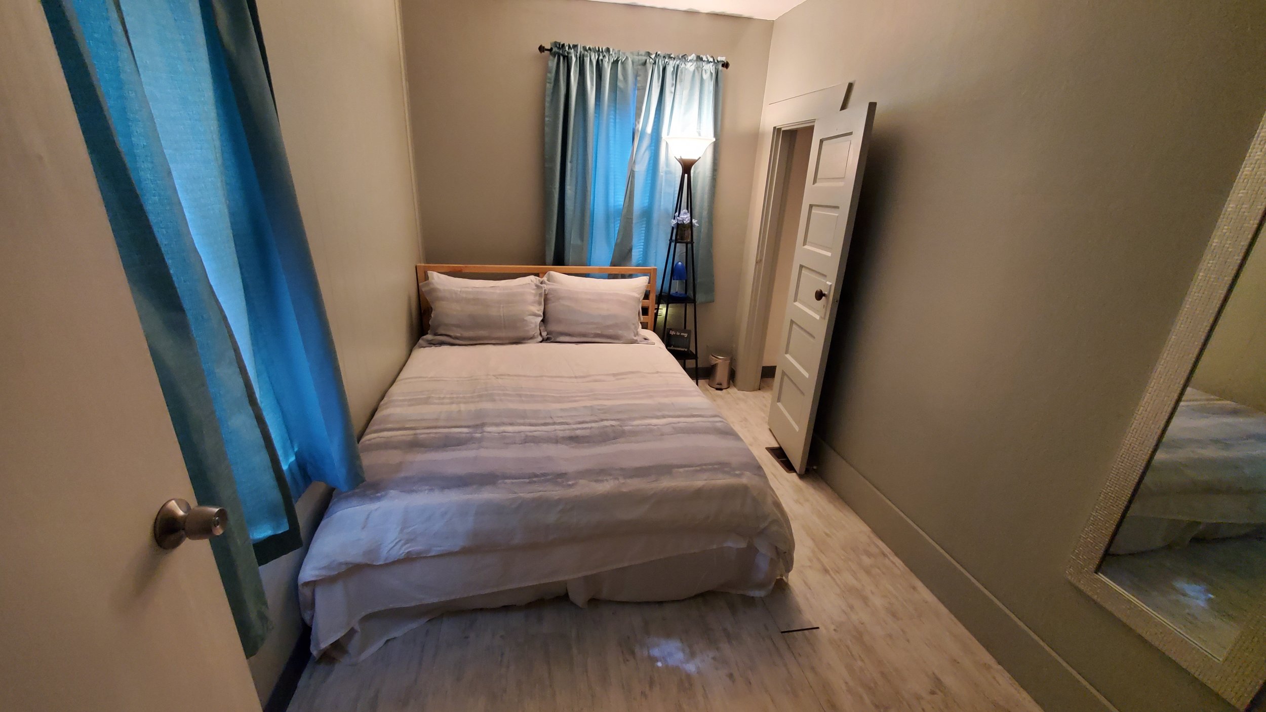 Small bedroom with a bed, two pillows, a standing light, window with blue curtains, and a large mirror on the wall.