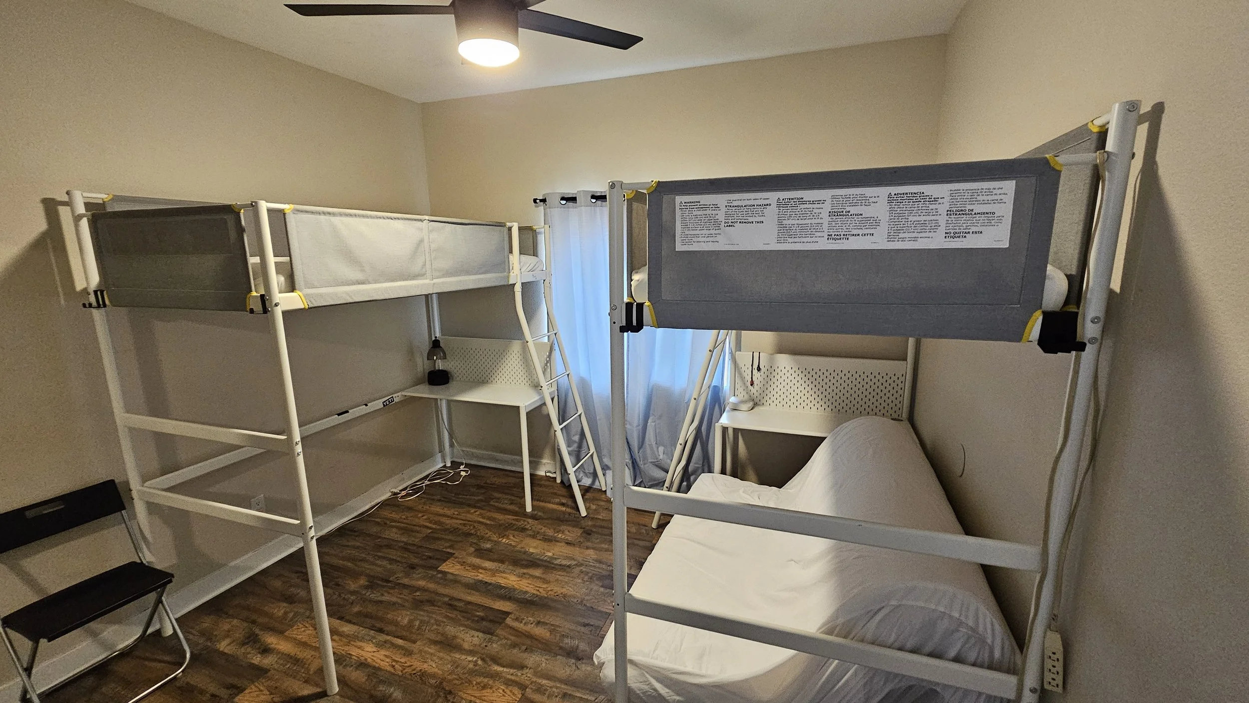 Room with two metal bunk beds, one with a mattress and pillow, a small white desk between the beds with a lamp, and a window with curtains. The floor is wood, and there is a ceiling fan with a light.