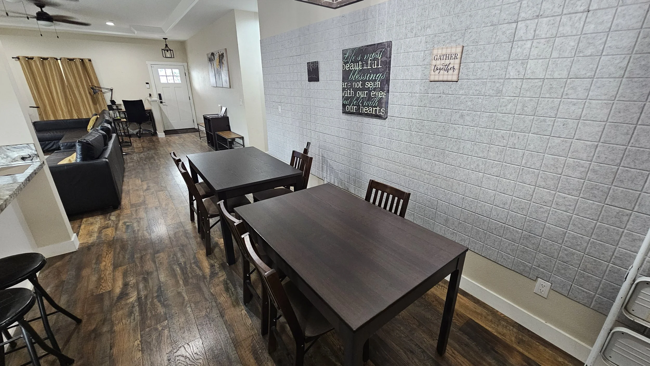 Dining area with two dark wooden tables, six chairs, a living area with black sofas, a white brick wall with decorative signs, hardwood floors, and a small corner desk near a window with gold curtains.