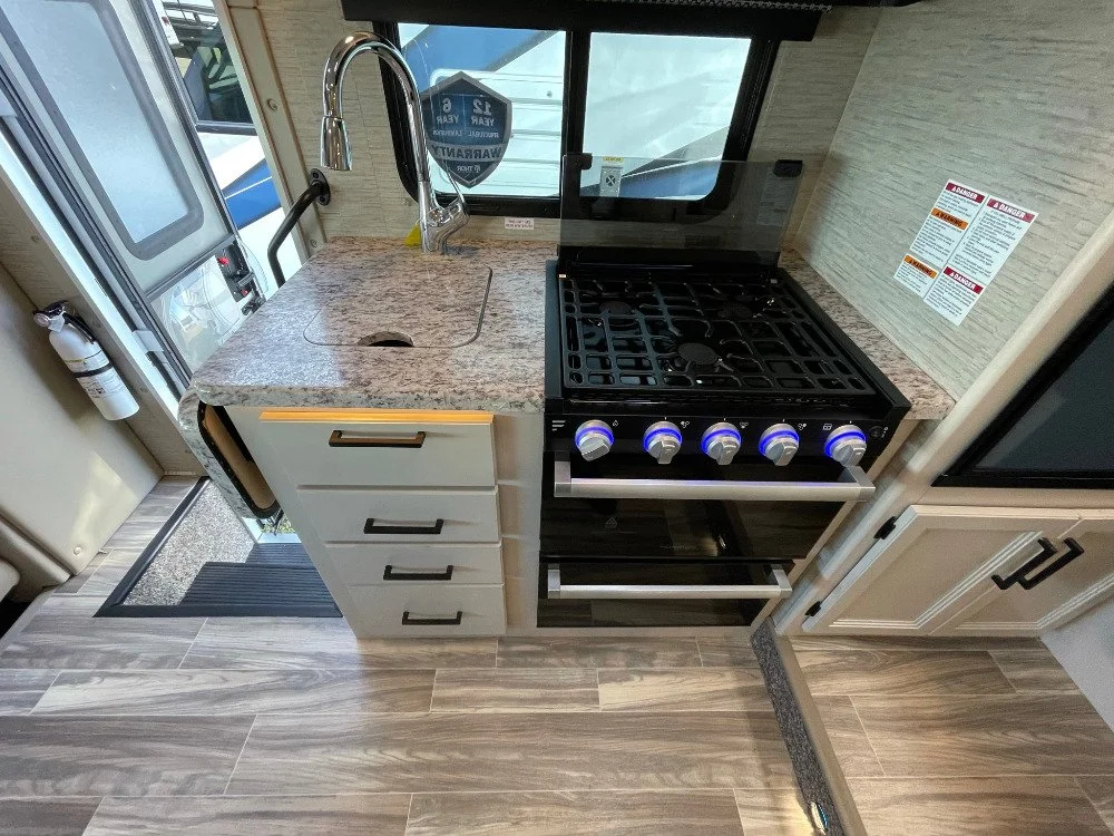 A compact kitchen area in an RV or camper with a granite countertop, a stainless steel sink with a curved faucet, a black stove with four silver knobs, a window behind the sink, and storage drawers and cabinets around.