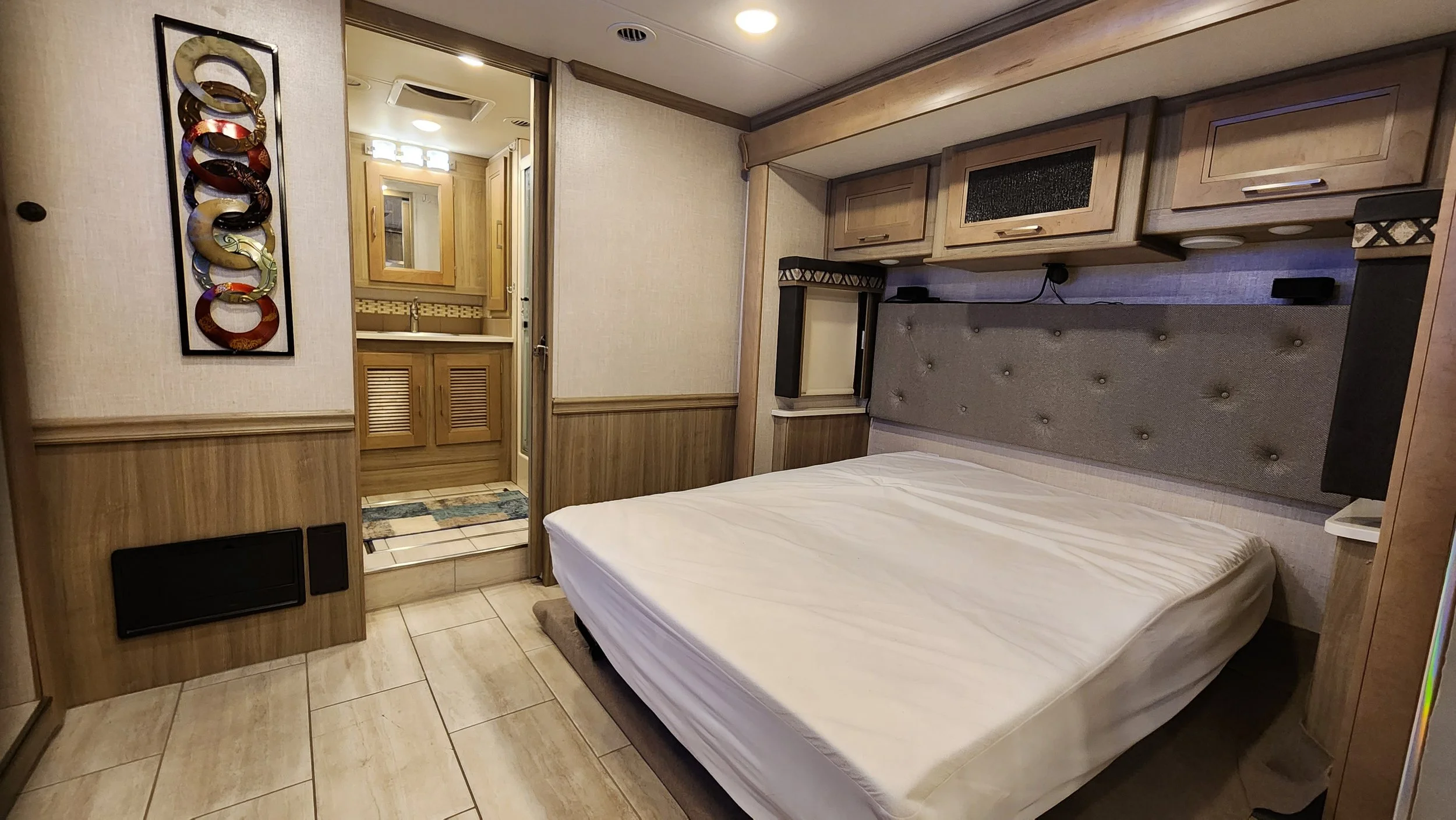 A bedroom inside a recreational vehicle or small living space with a bed, overhead wooden cabinets, and a view into a bathroom with a sink and mirror.