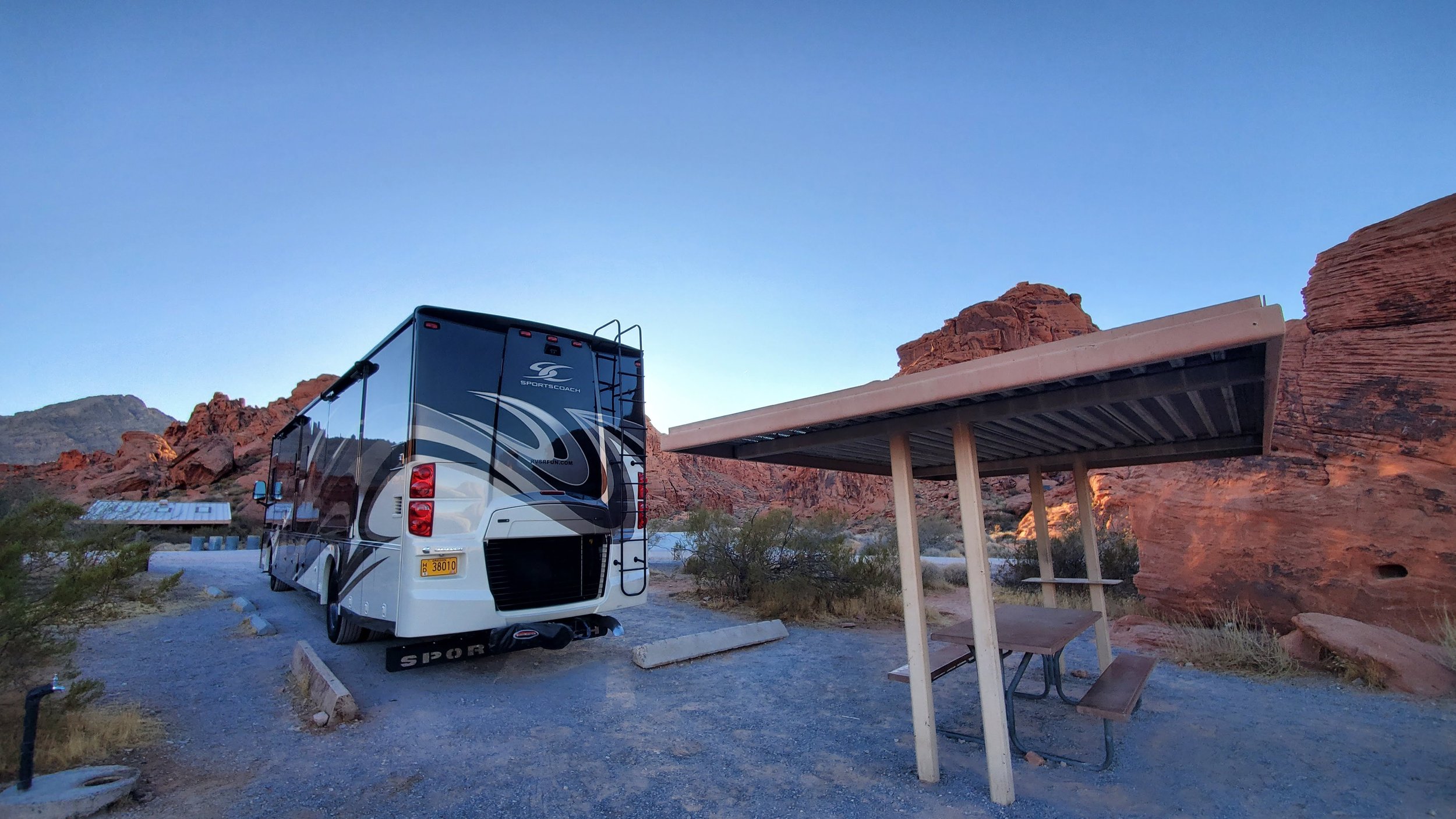 A parked recreational vehicle next to a picnic shelter in a desert landscape with red rock formations and mountains in the background at dusk.