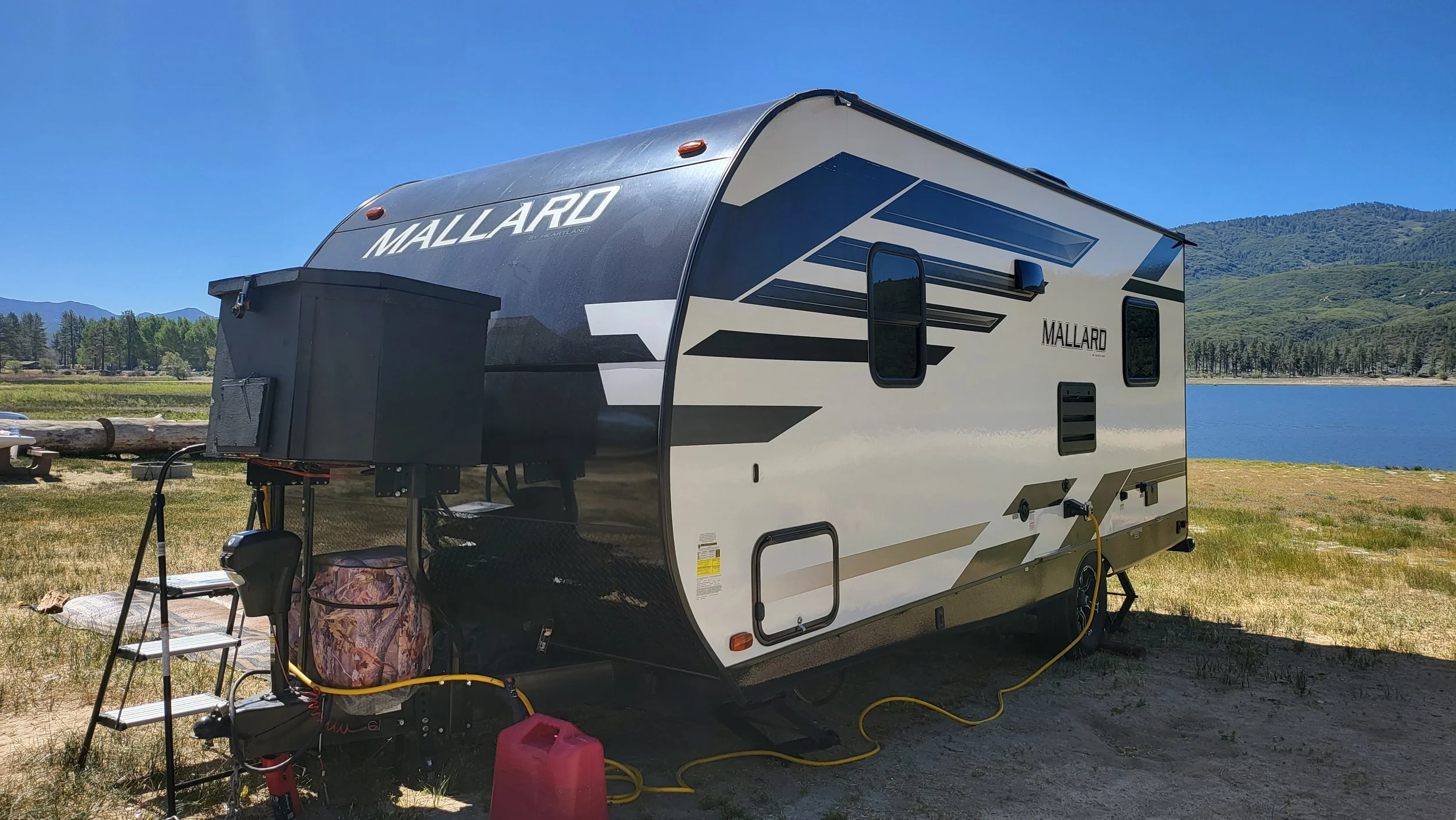 Black and white Mallard travel trailer parked by a lake with mountains in the background, connected to power and water supplies.