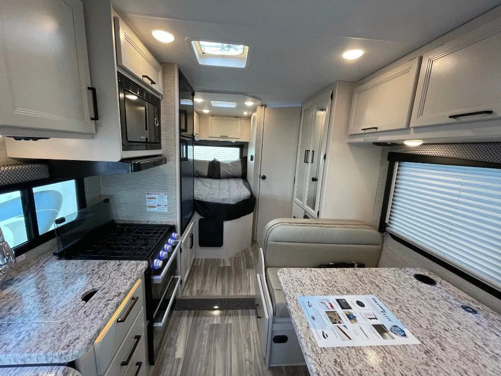 Interior of an RV with a kitchen area featuring a stove, microwave, and countertop, a dining table, and a sleeping area with a bed at the back.