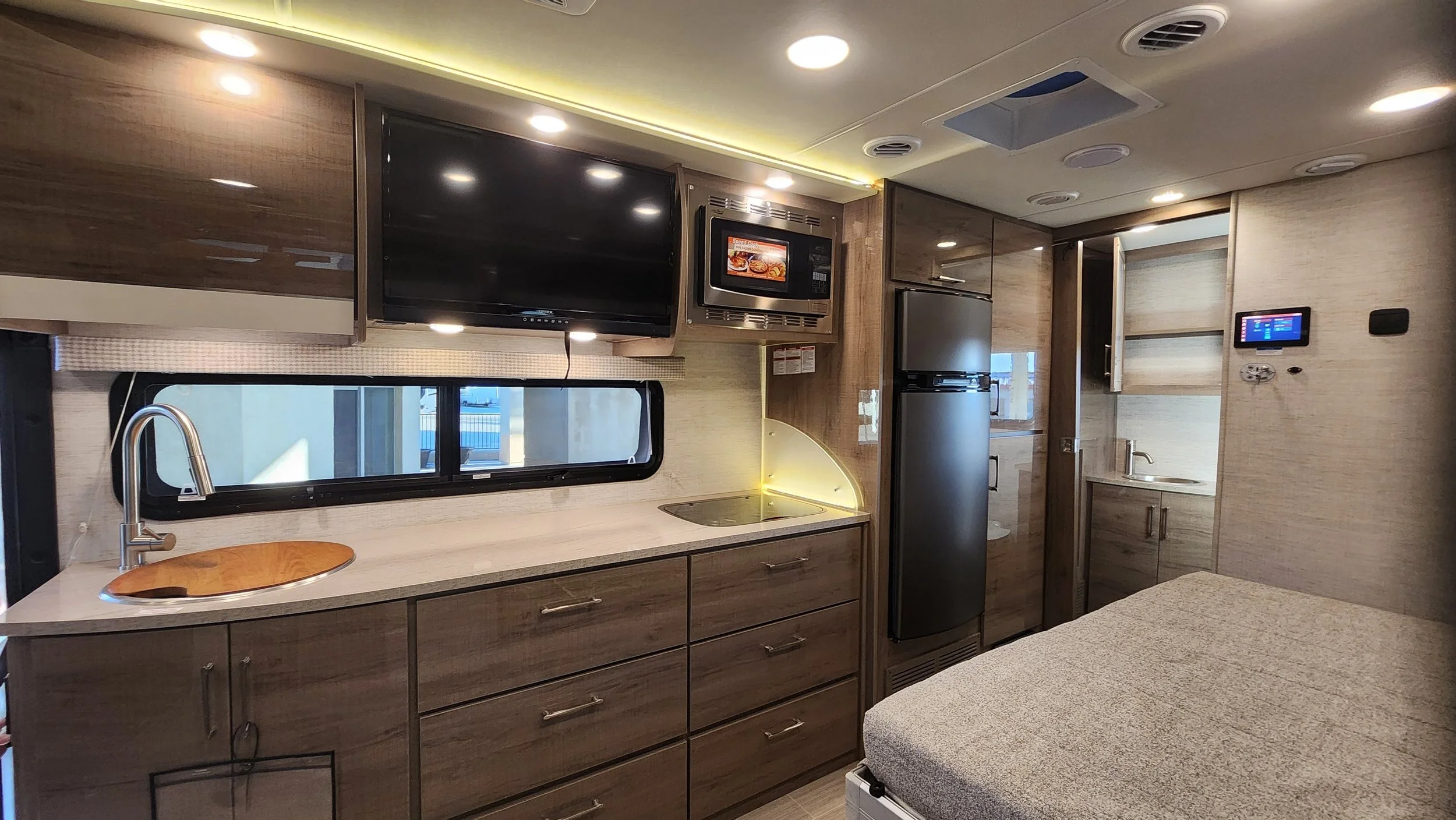 Interior of a modern RV or camper kitchen with wood cabinets, a small sink, microwave, TV, refrigerator, and a bed nearby.