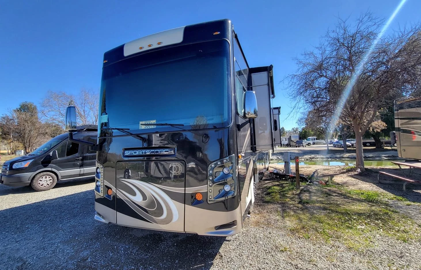 Black and beige Sportscoach RV parked on gravel near trees and a pond with a clear blue sky.