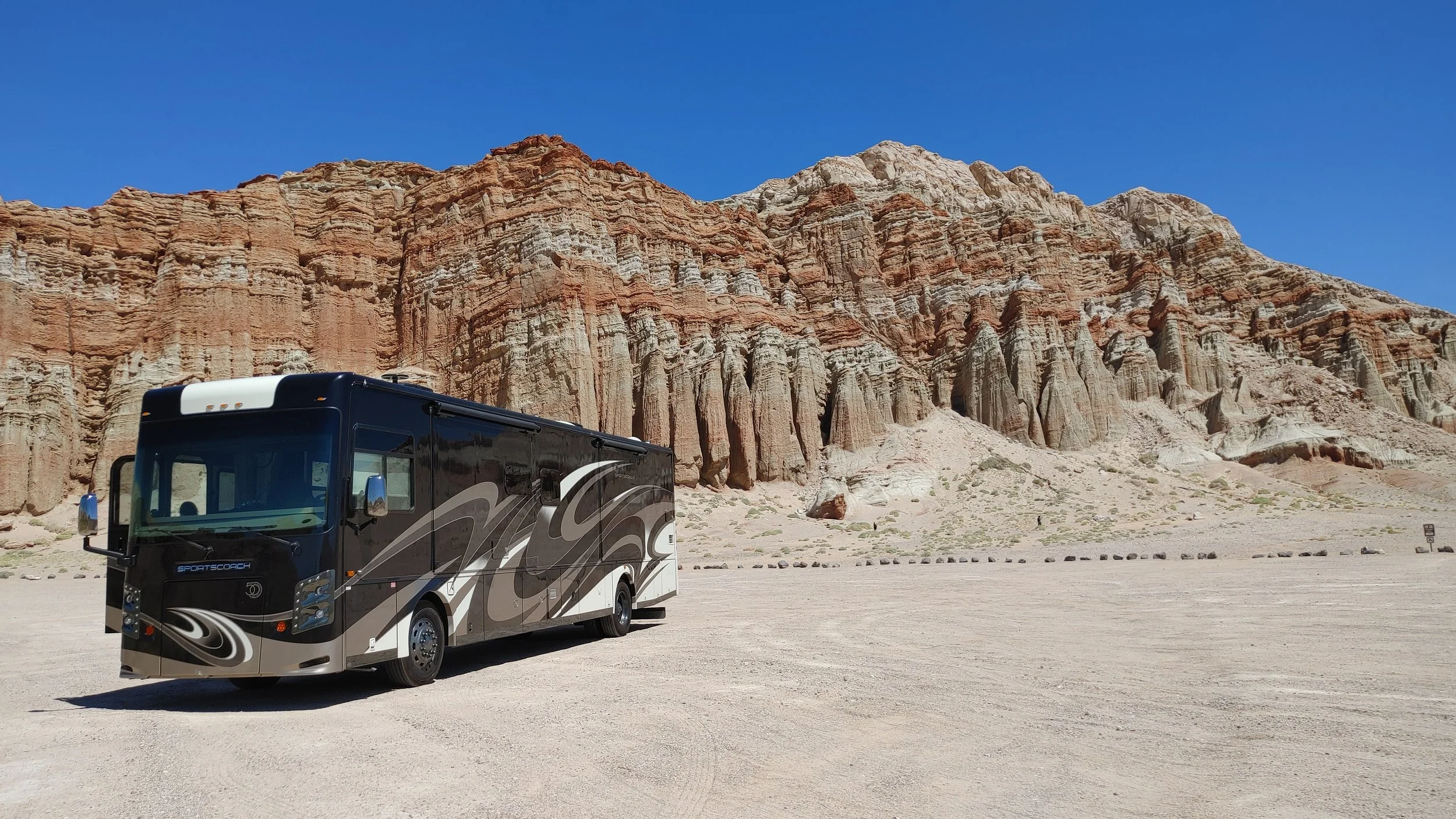 A black and white recreational vehicle parked on a dirt area with a rocky cliff in the background under a clear blue sky.