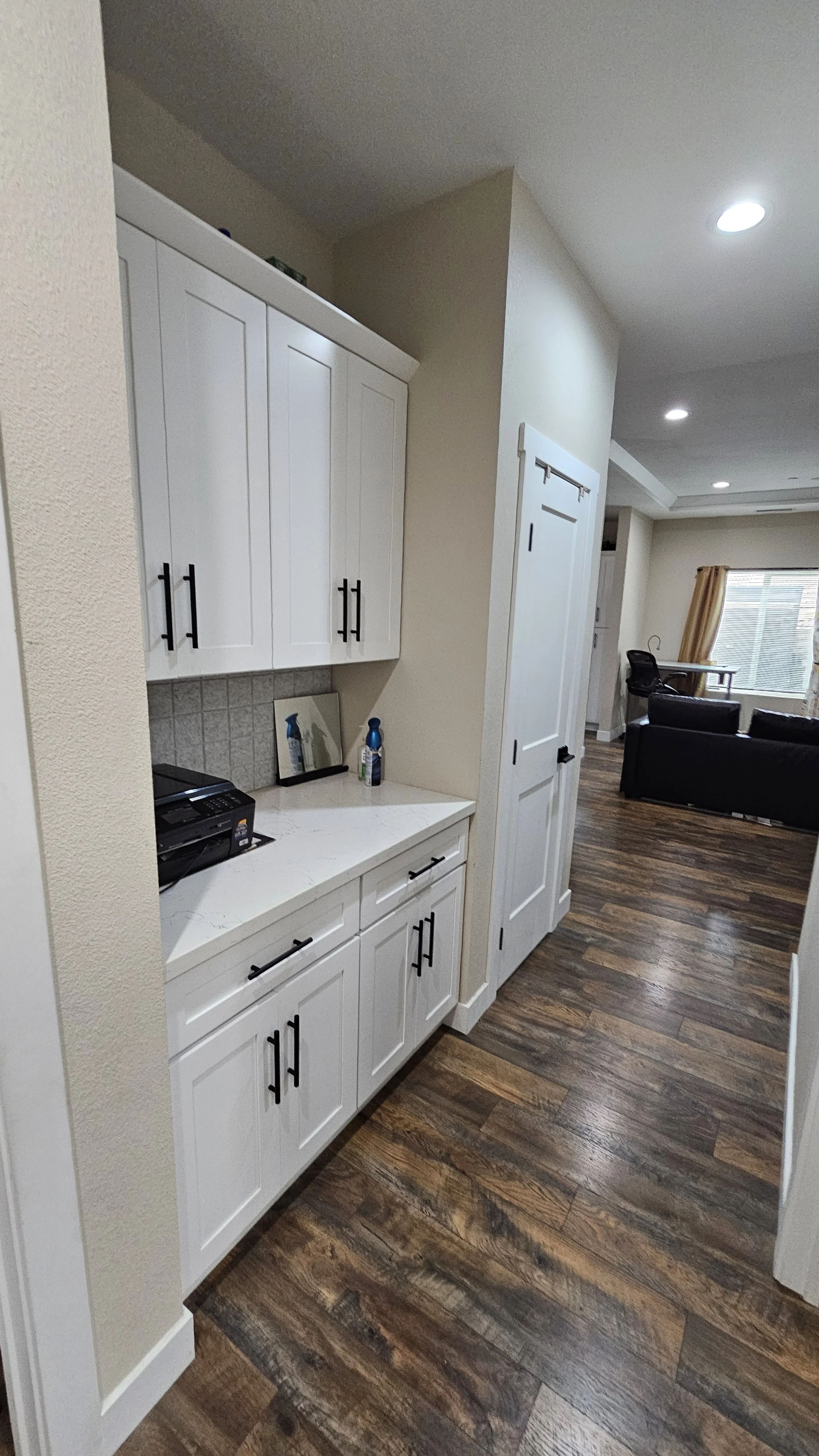 Kitchen corner with white cabinets, black handles, a countertop with a printer, cleaning spray, mirror, and door, overlooking a living room with a window, curtains, and furniture.