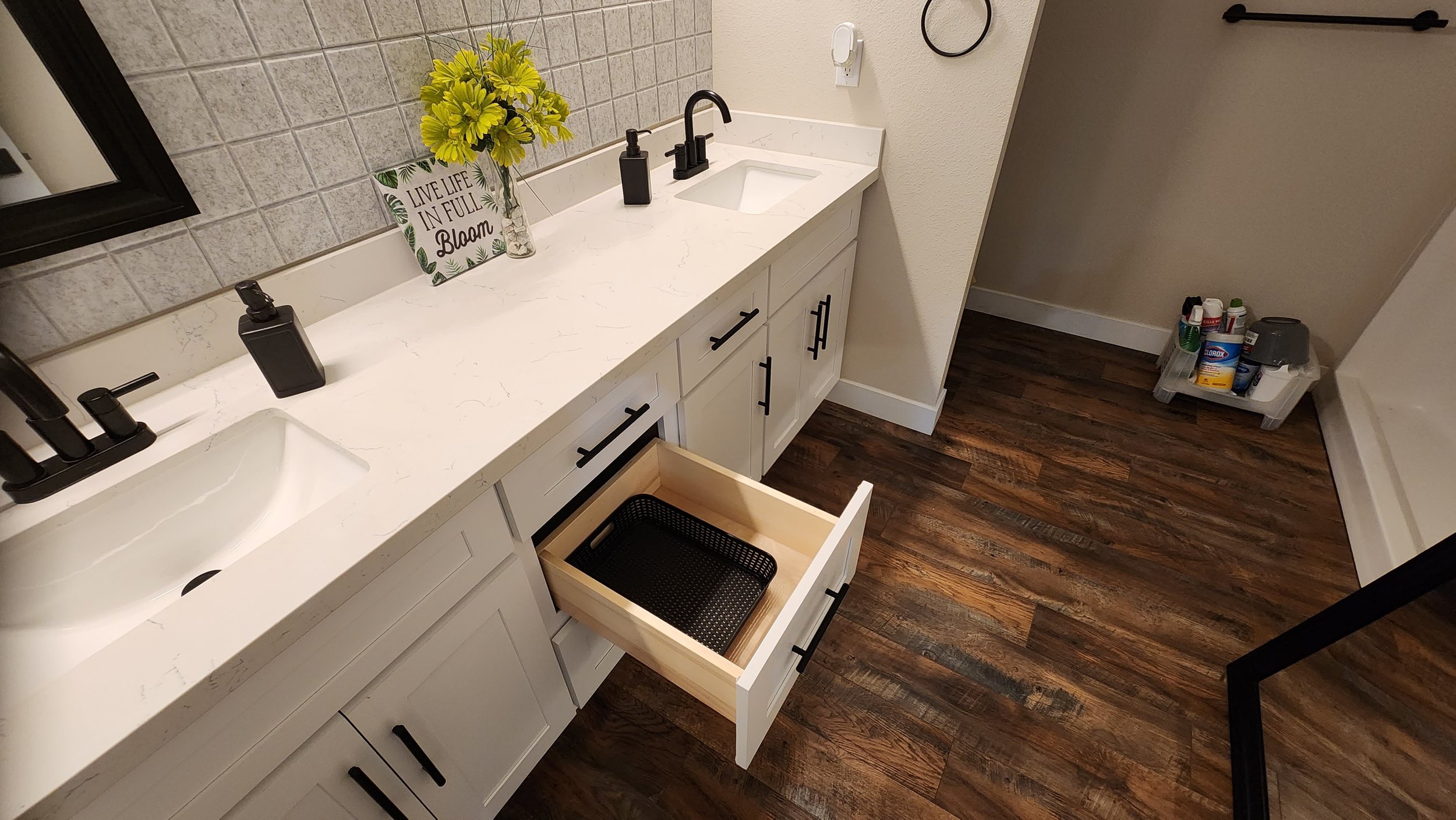 Bathroom vanity with two sinks, a mirror, a soap dispenser, a flower arrangement, a sign reading 'Live Life in Full Bloom,' and open drawers showing storage space. There is a small storage cart with cleaning supplies and a wood-floor flooring.