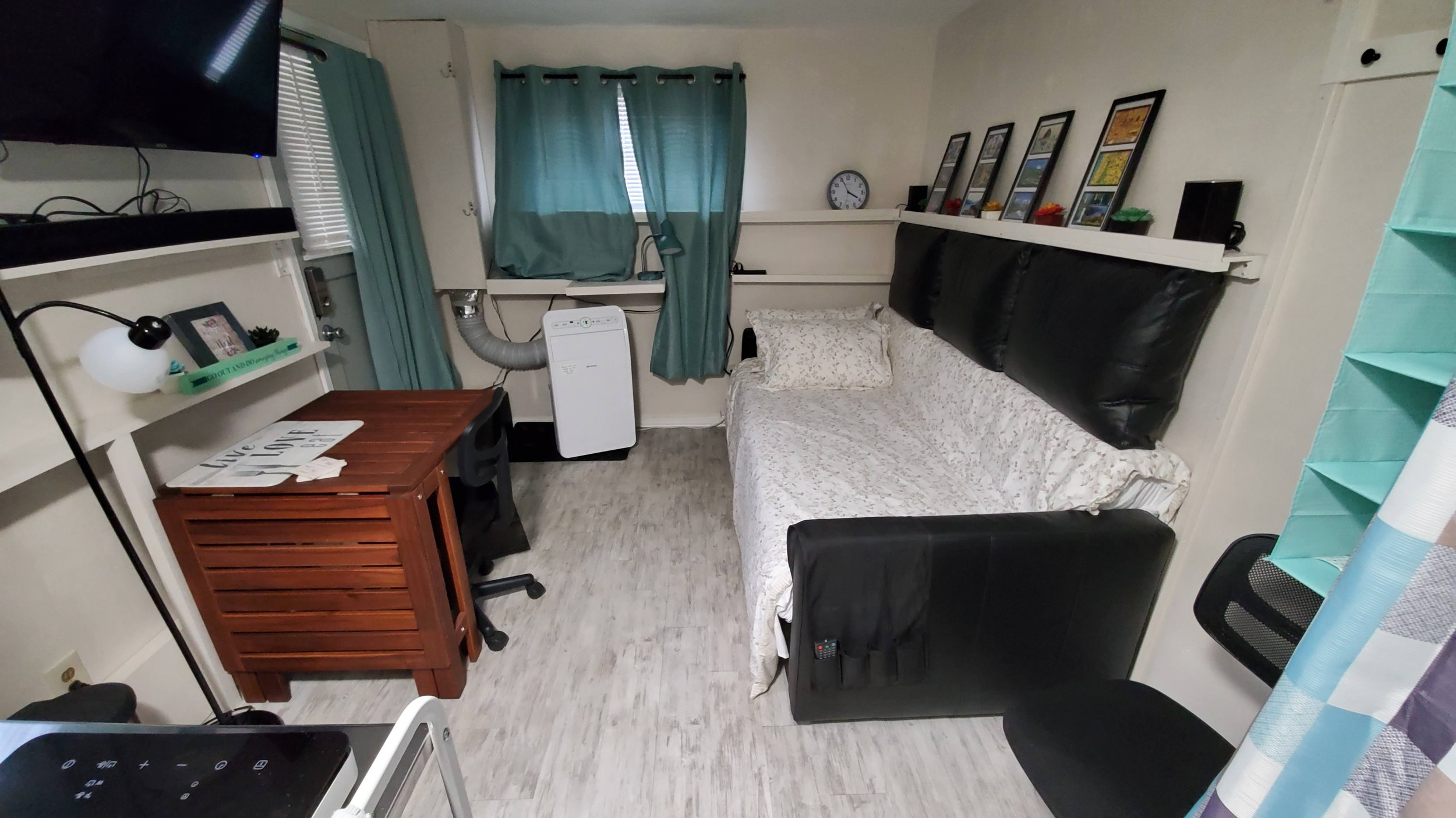 A small room with a couch, framed pictures on a shelf, a window with blue curtains, a wooden desk, a chair, a lamp, a small air purifier, and a TV mounted on the wall.