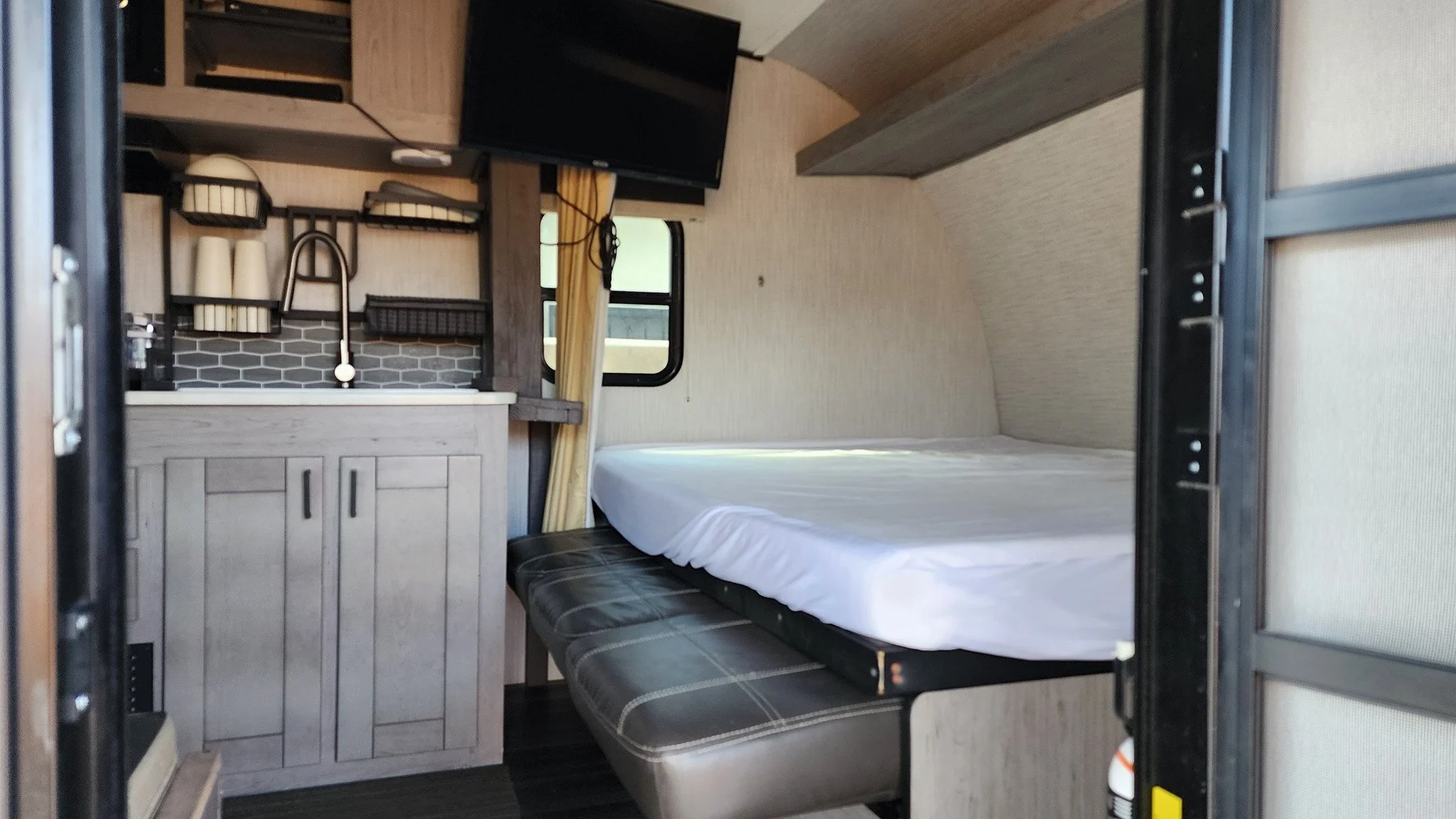 Interior of a compact camper or tiny home with a small kitchen, a bed, and a television, featuring wood finish and minimal decor.