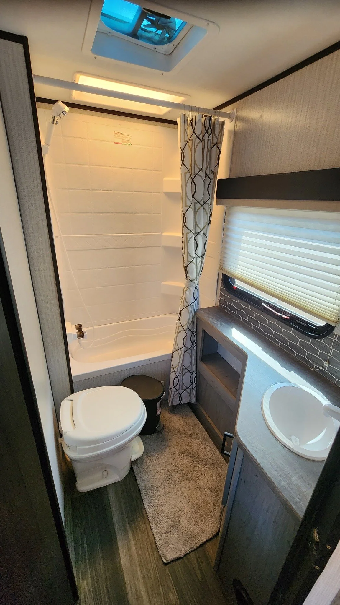 Small bathroom with a toilet, a shower with a curtain, a small sink, a window with blinds, and a skylight on the ceiling.