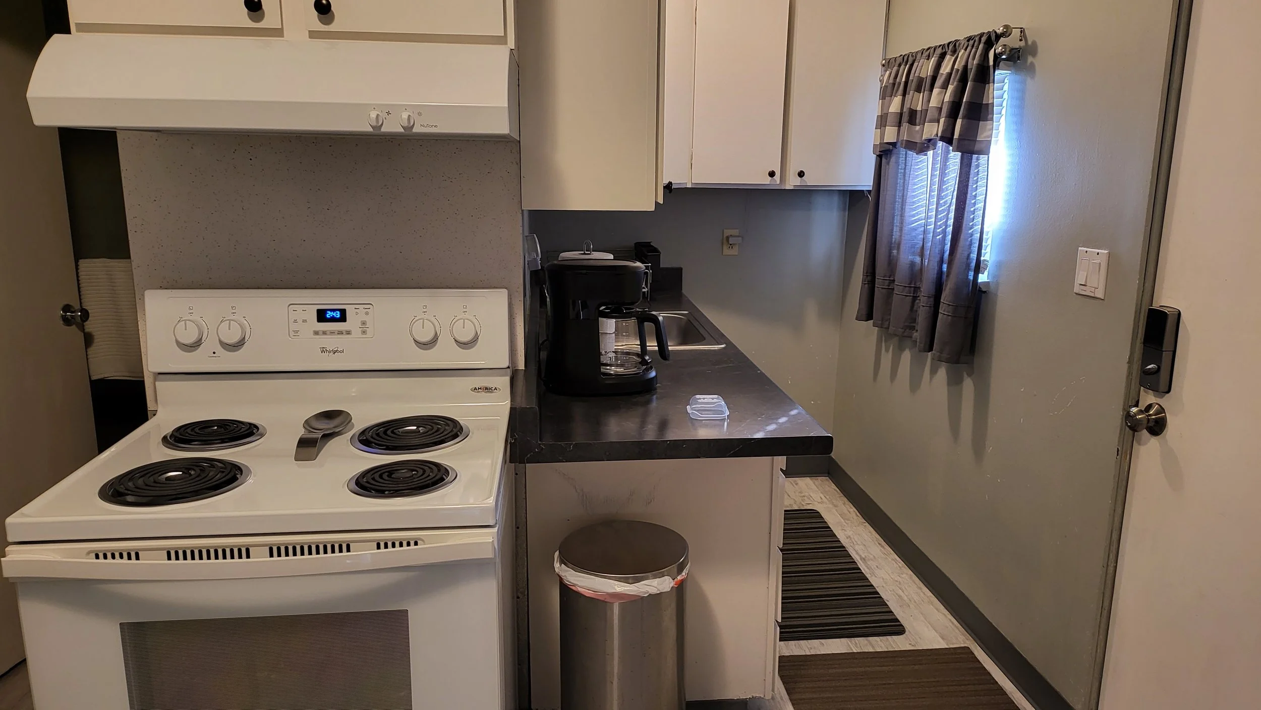 A small kitchen with a white stove, black coffee maker, white cabinets, and a small window with checkered curtains.