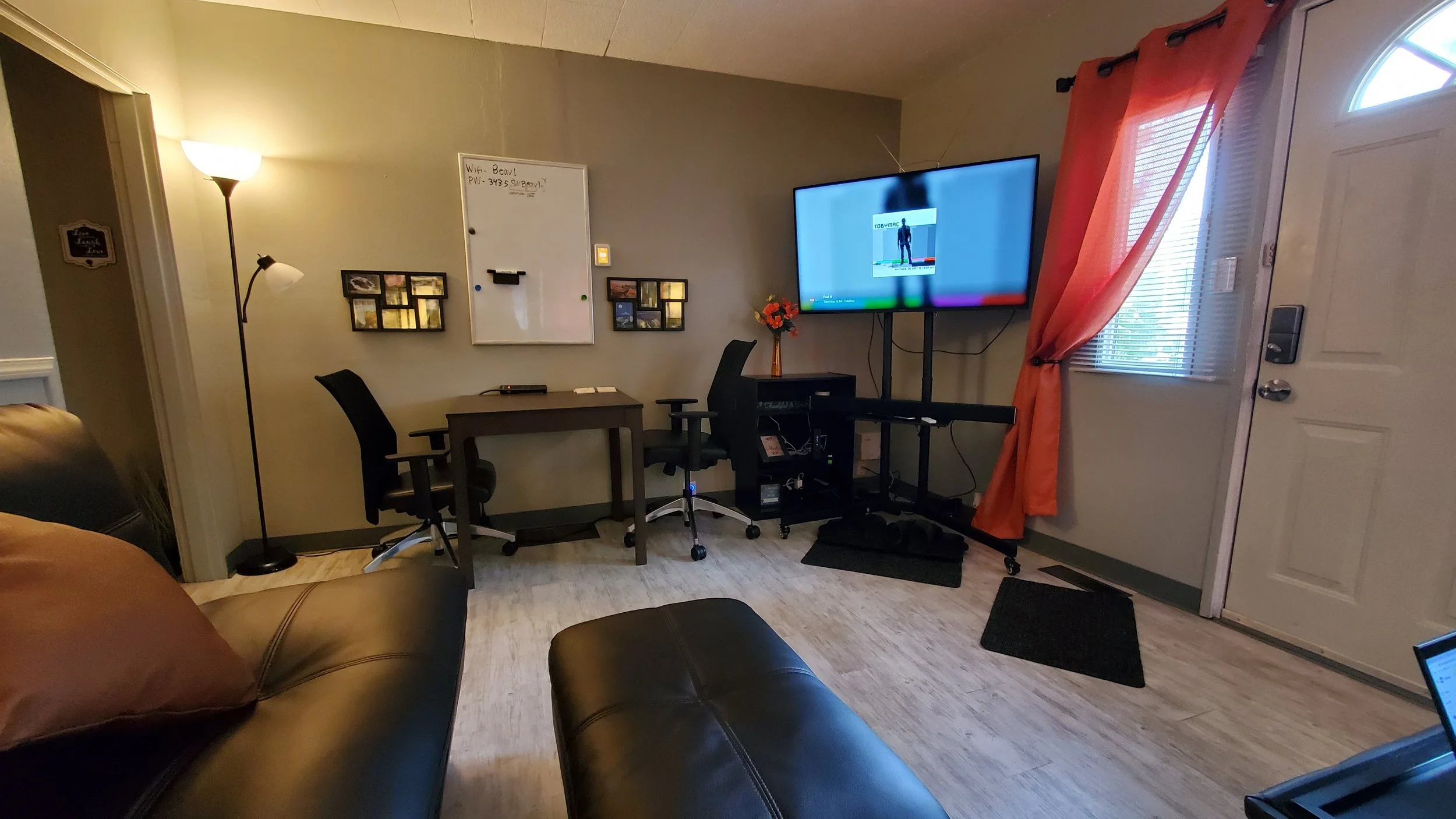 Living room with a leather sofa, a standing lamp, a small table with two office chairs, a wall whiteboard, and a large wall-mounted television. There are orange curtains over a window/door and a small black mat on the floor.