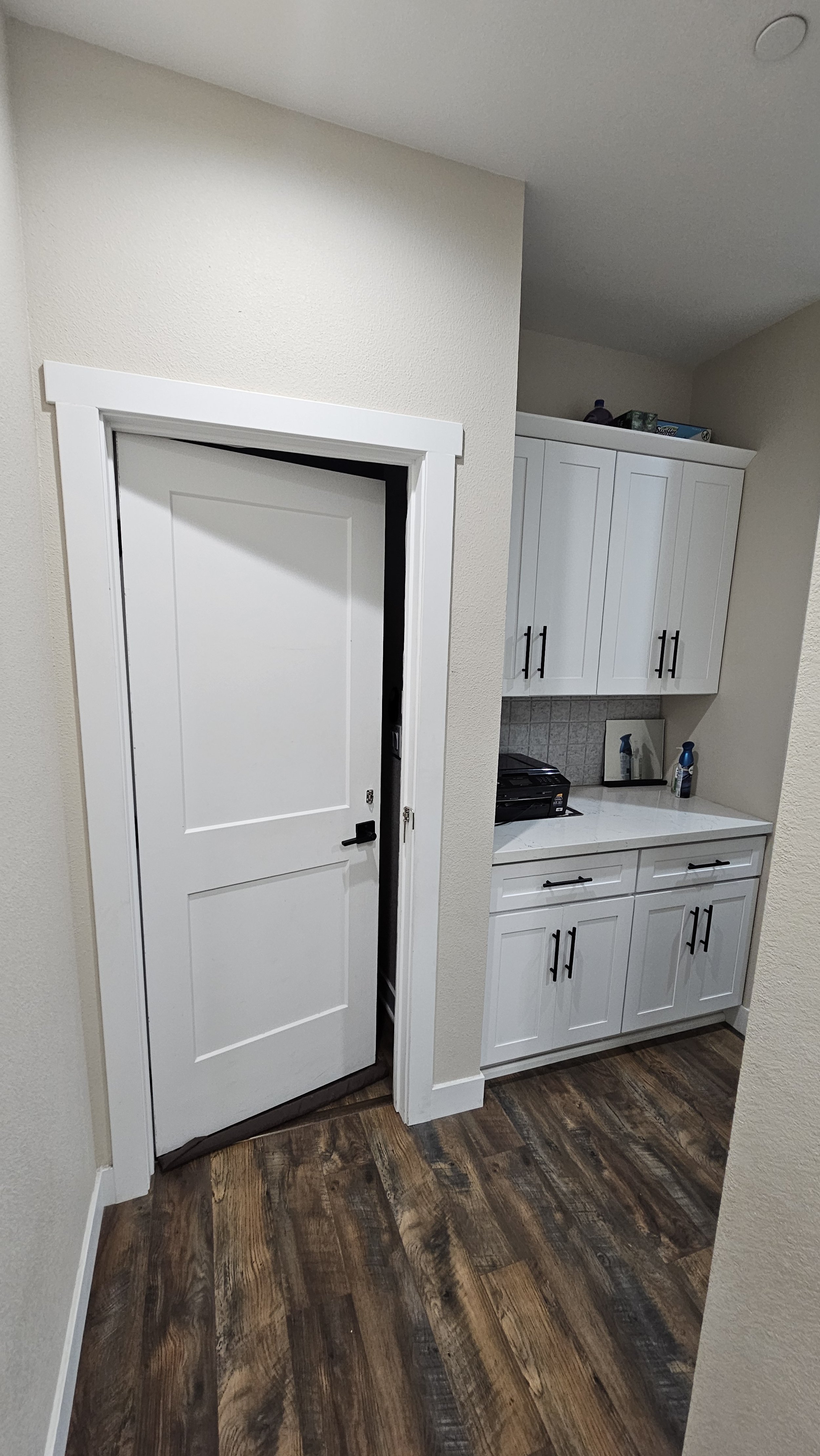 A white interior door slightly ajar, adjacent to a white kitchen cabinet with black handles, on a wood floor with a dark stain.