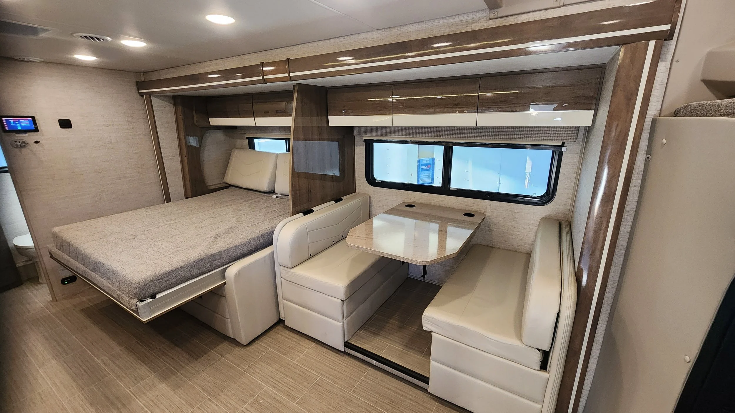 Interior of a motorhome or camper with a bed, dining table, and seating area, featuring wood and cream-colored upholstery, windows, and ceiling lights.