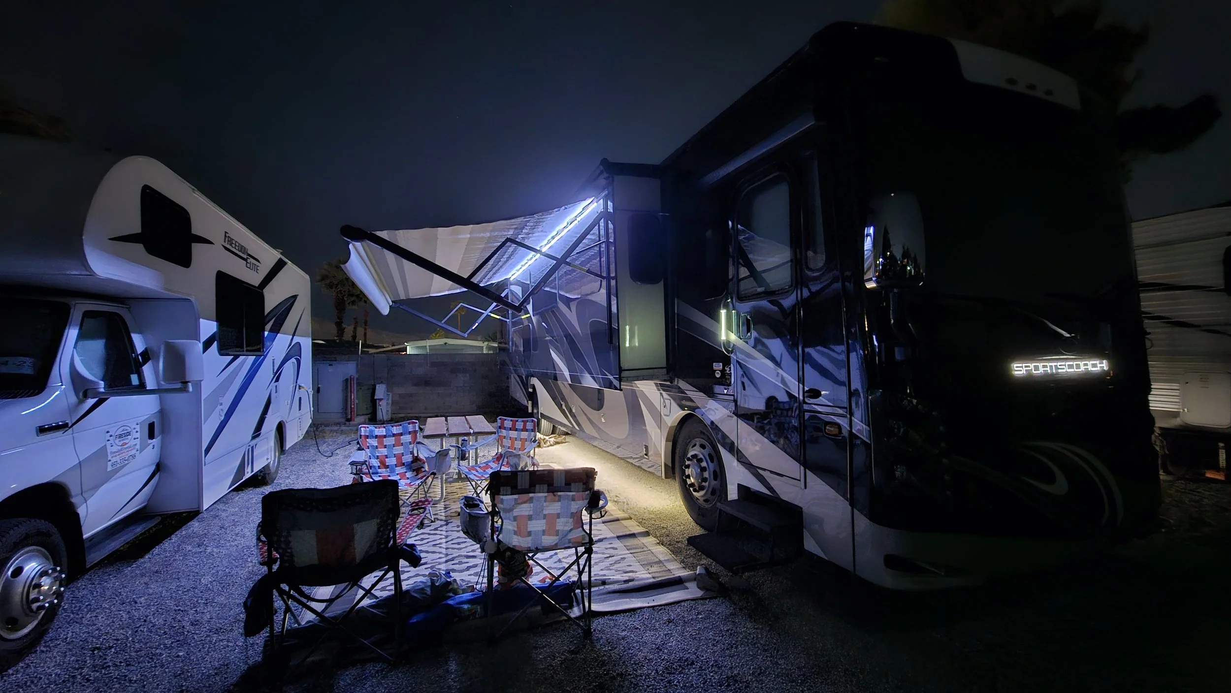 Nighttime scene with two recreational vehicles, one with an extended awning, outdoor chairs, and a rug on gravel ground.
