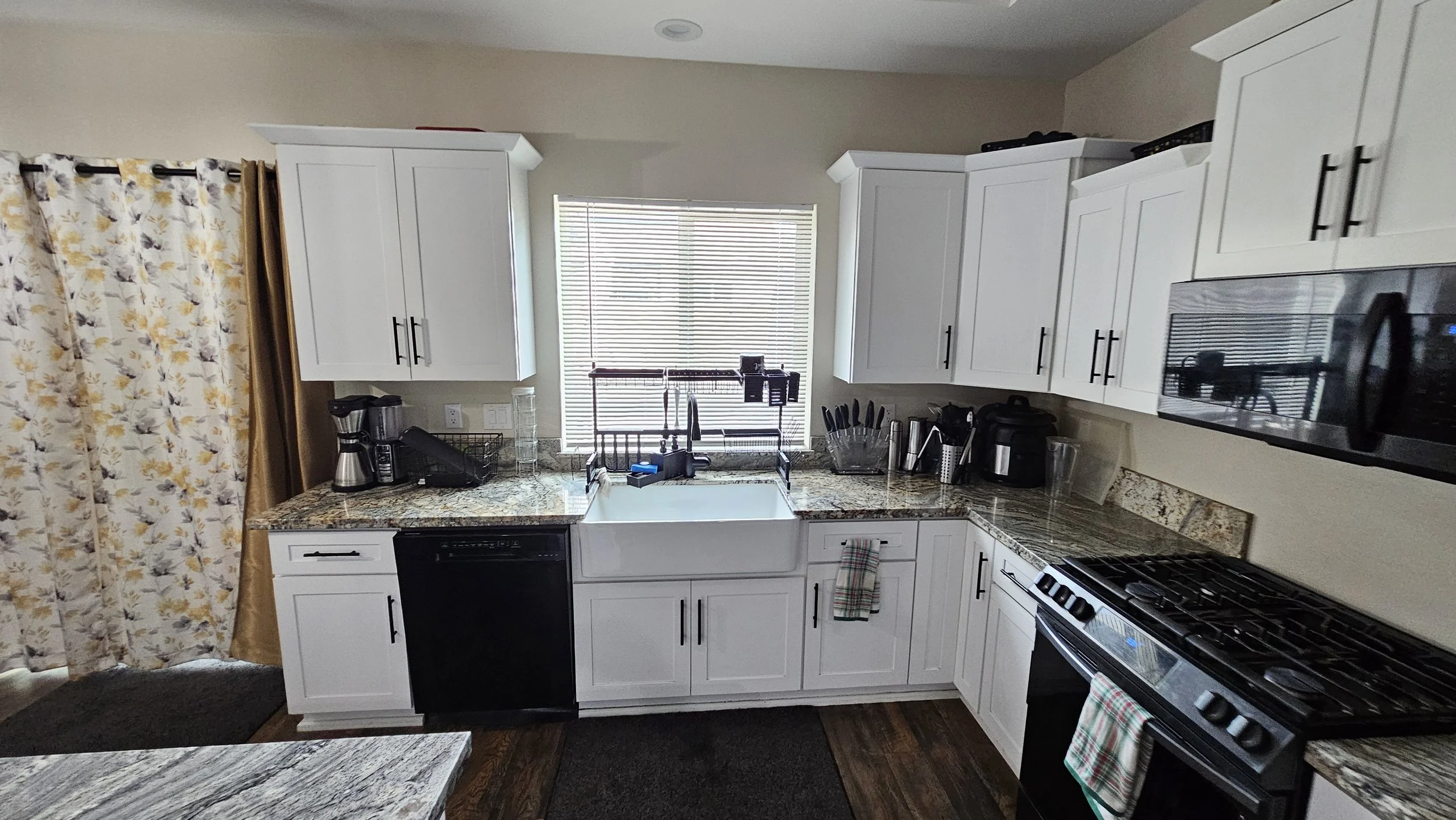 Kitchen with white cabinets, granite countertops, a window above the sink, a dishwasher, a stove, microwave, coffee maker, kitchen utensils, and curtains with a floral pattern.